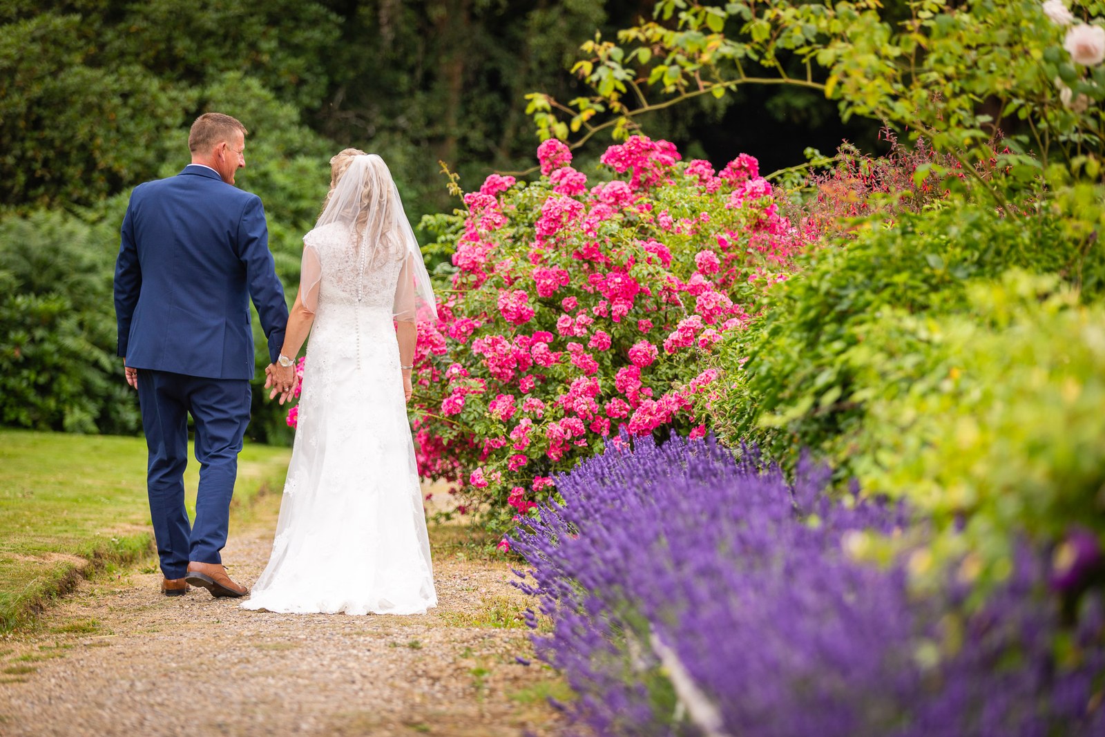 The couple walking a rose-and-lavender garden path at Grim's Dyke Hotel, seen from behind