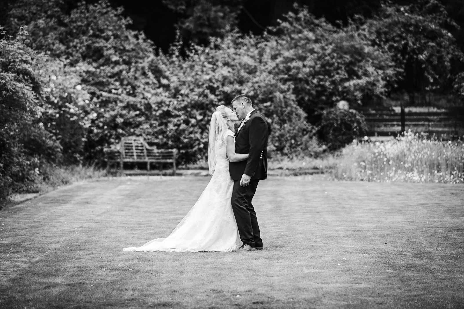 Black-and-white of the couple kissing on a wide lawn framed by trees at Grim's Dyke Hotel