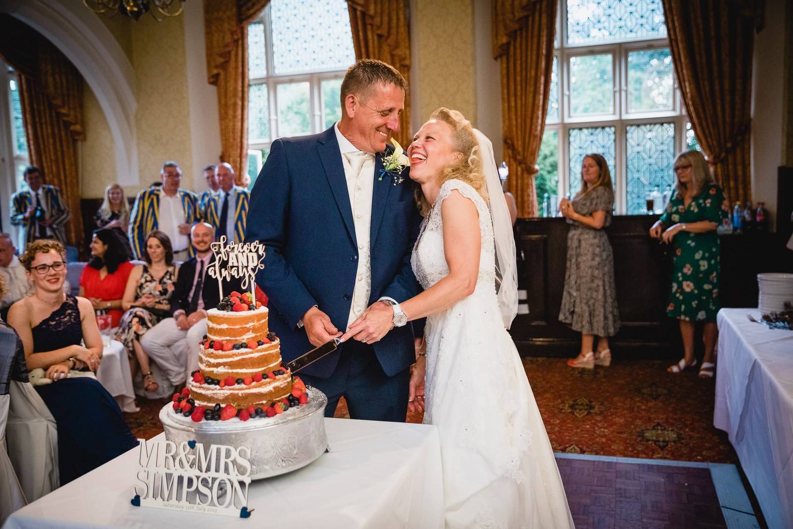 The couple cutting their berry-topped naked cake with a personalised topper as guests watch