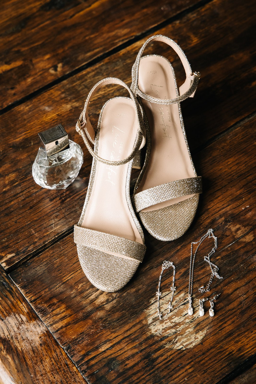 Sparkly gold heeled sandals beside a glass perfume bottle and drop earrings on a dark wooden floor