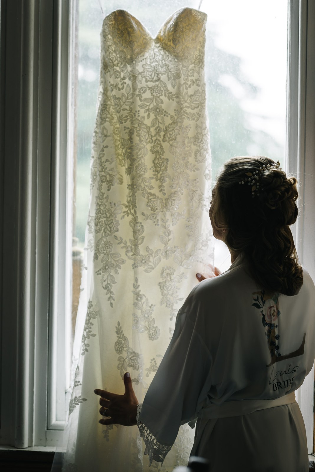 Bride in a robe gazing at her lace gown hanging in the window light