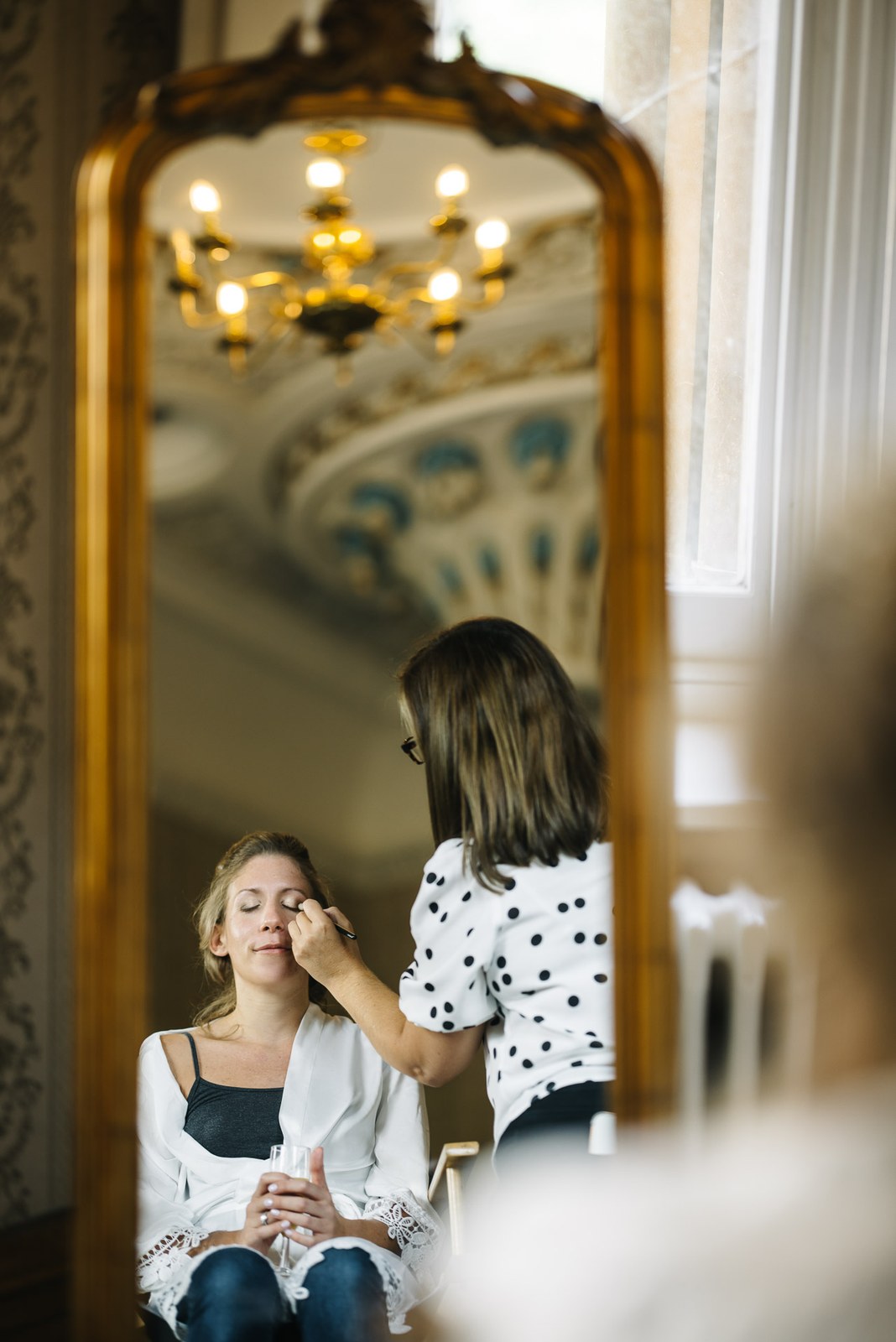 A woman having her make-up applied, reflected in an ornate gilt mirror