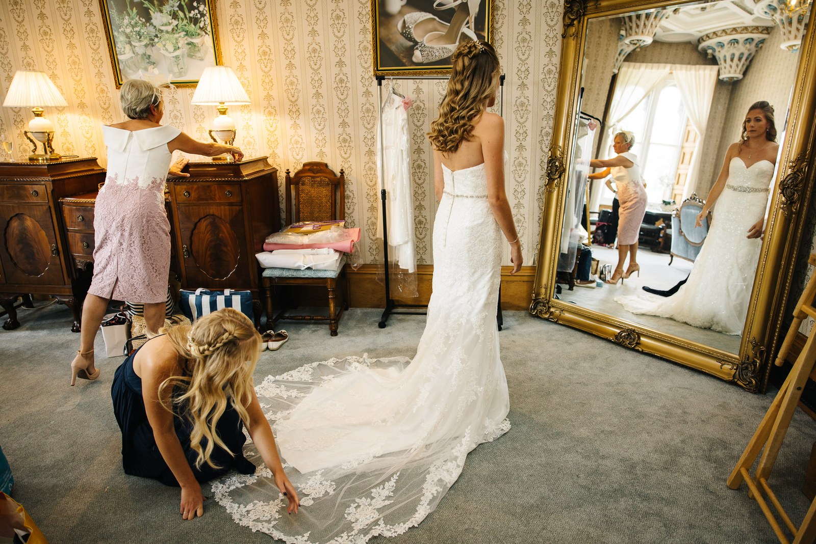 Bride in her gown before a tall gilt mirror as a bridesmaid lays out the train