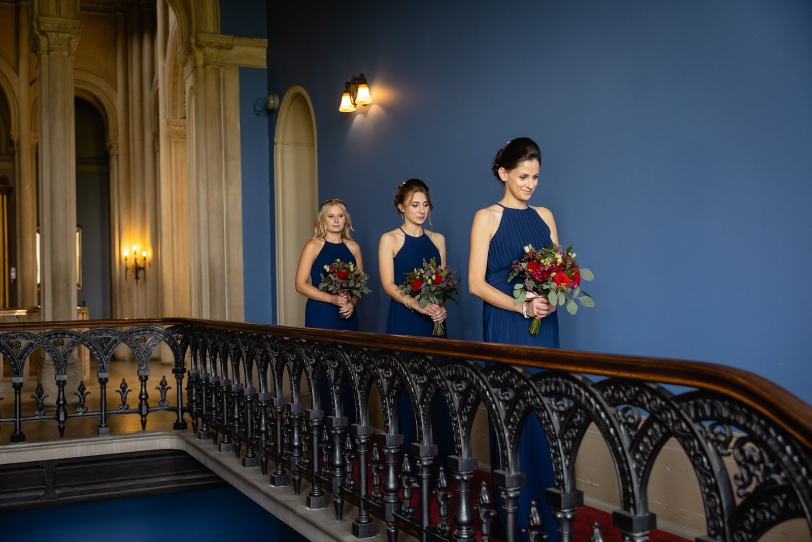 Bridesmaids in navy with red bouquets on the landing above the ornate ironwork staircase
