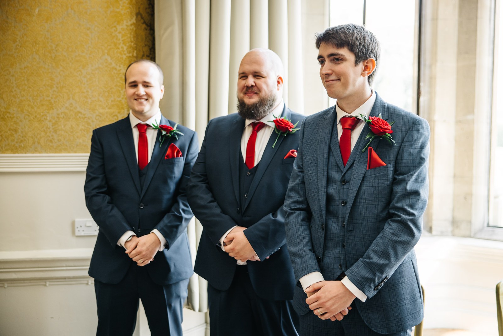 The groom and his two groomsmen in blue suits with red ties and rose buttonholes