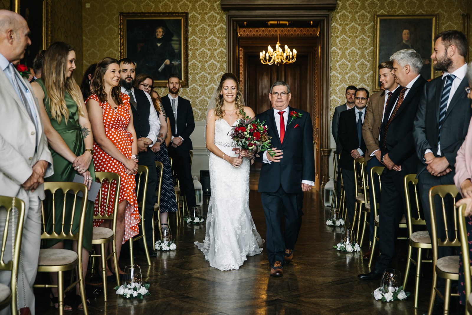 Bride on her father's arm entering the aisle as seated guests rise, damask ceremony room