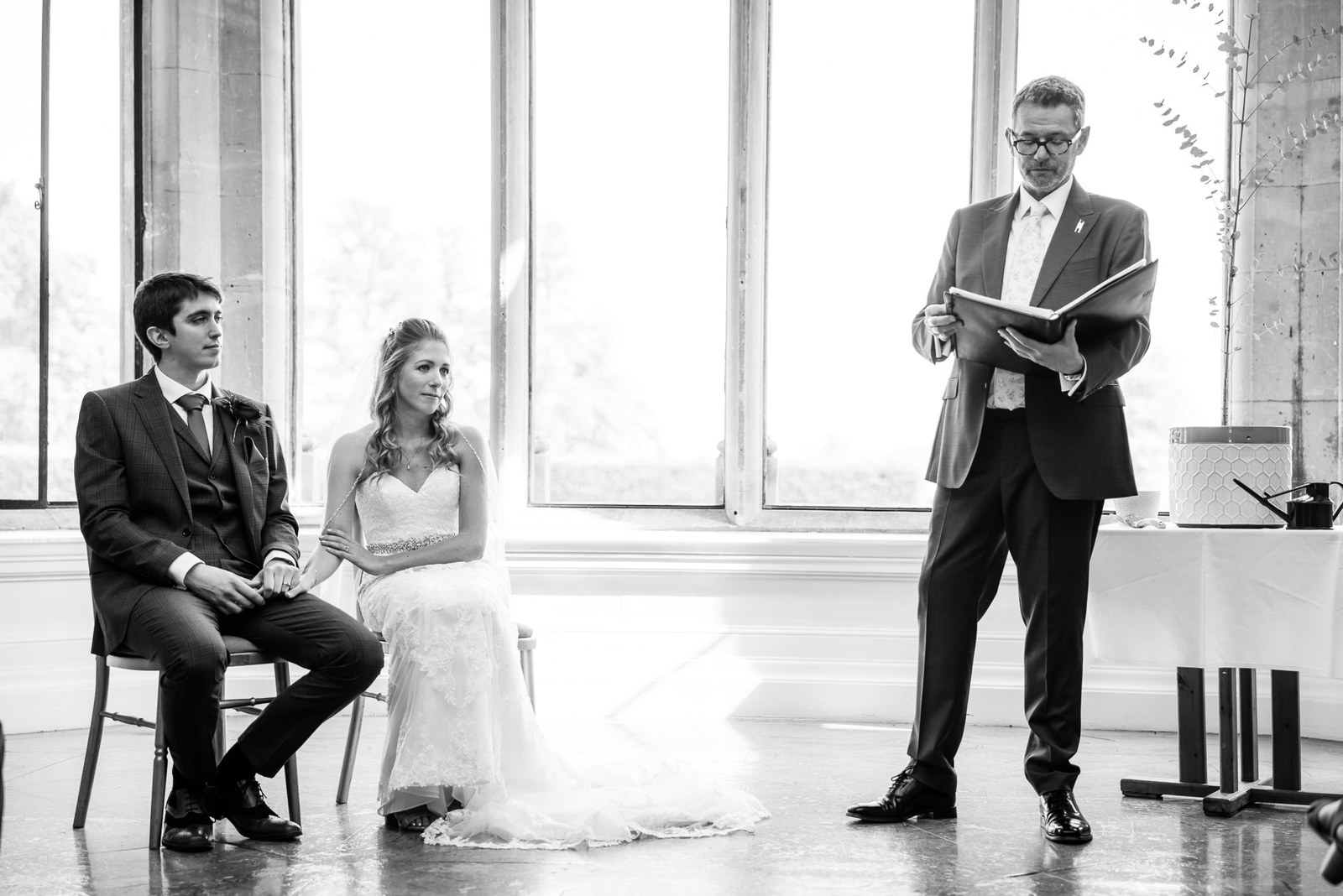 Black-and-white ceremony moment, the couple seated as the registrar reads by the window