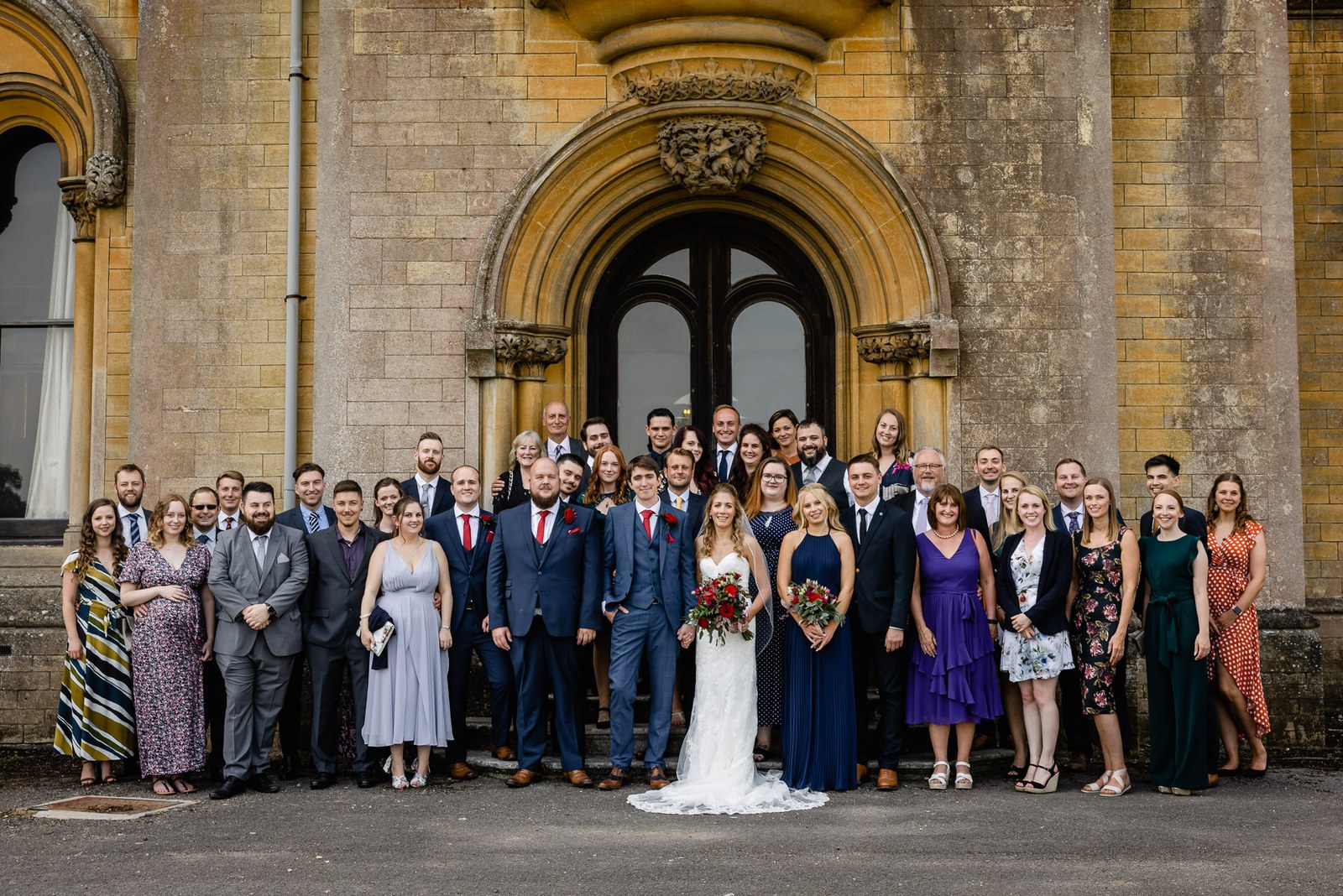 The full wedding group posed beneath the arched Gothic entrance of Grittleton House