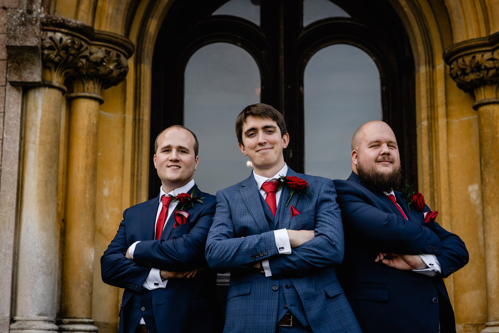 The groom and his two groomsmen with folded arms before the golden stone arches