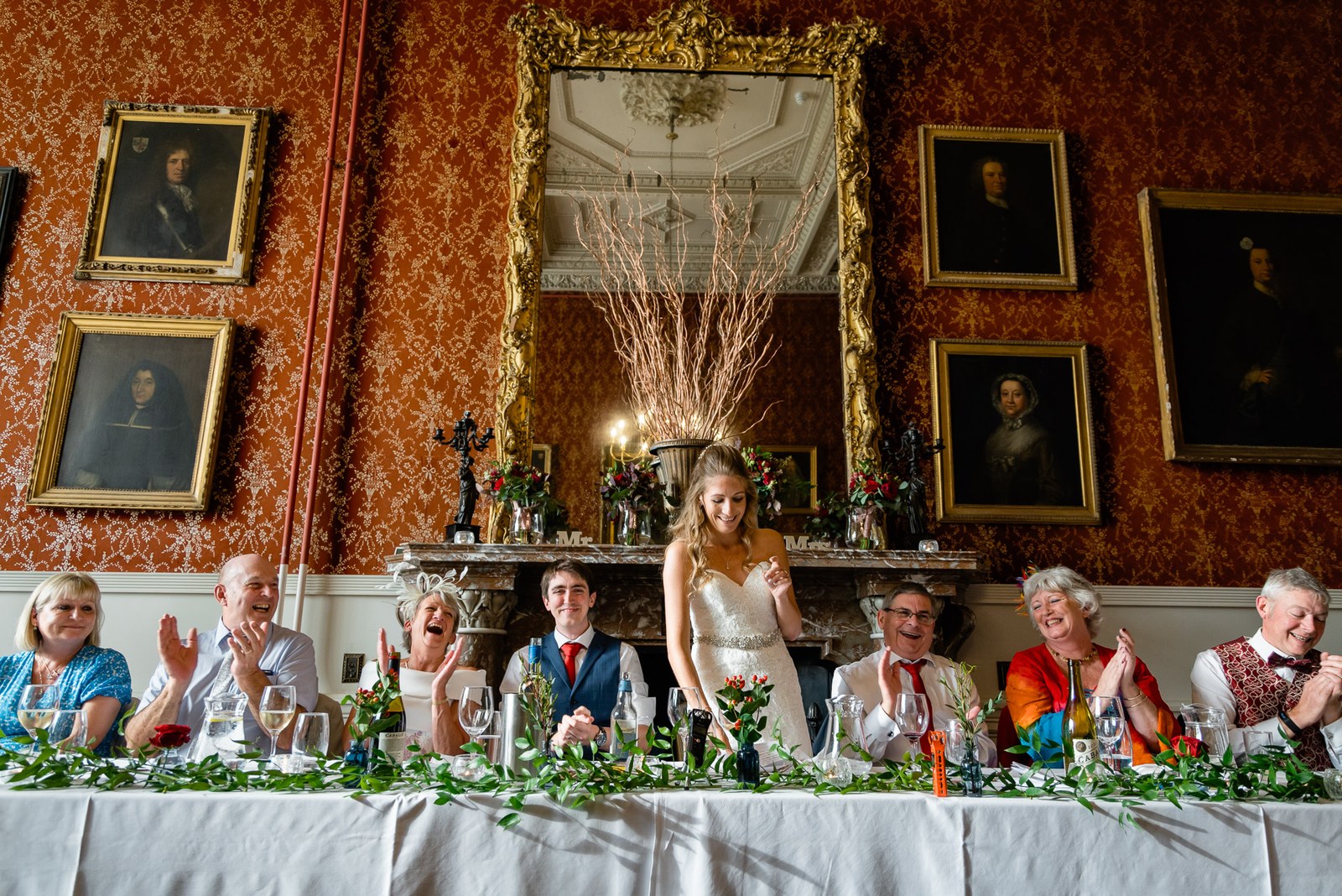 The bride standing at the top table to applause in the ornate red-walled dining room
