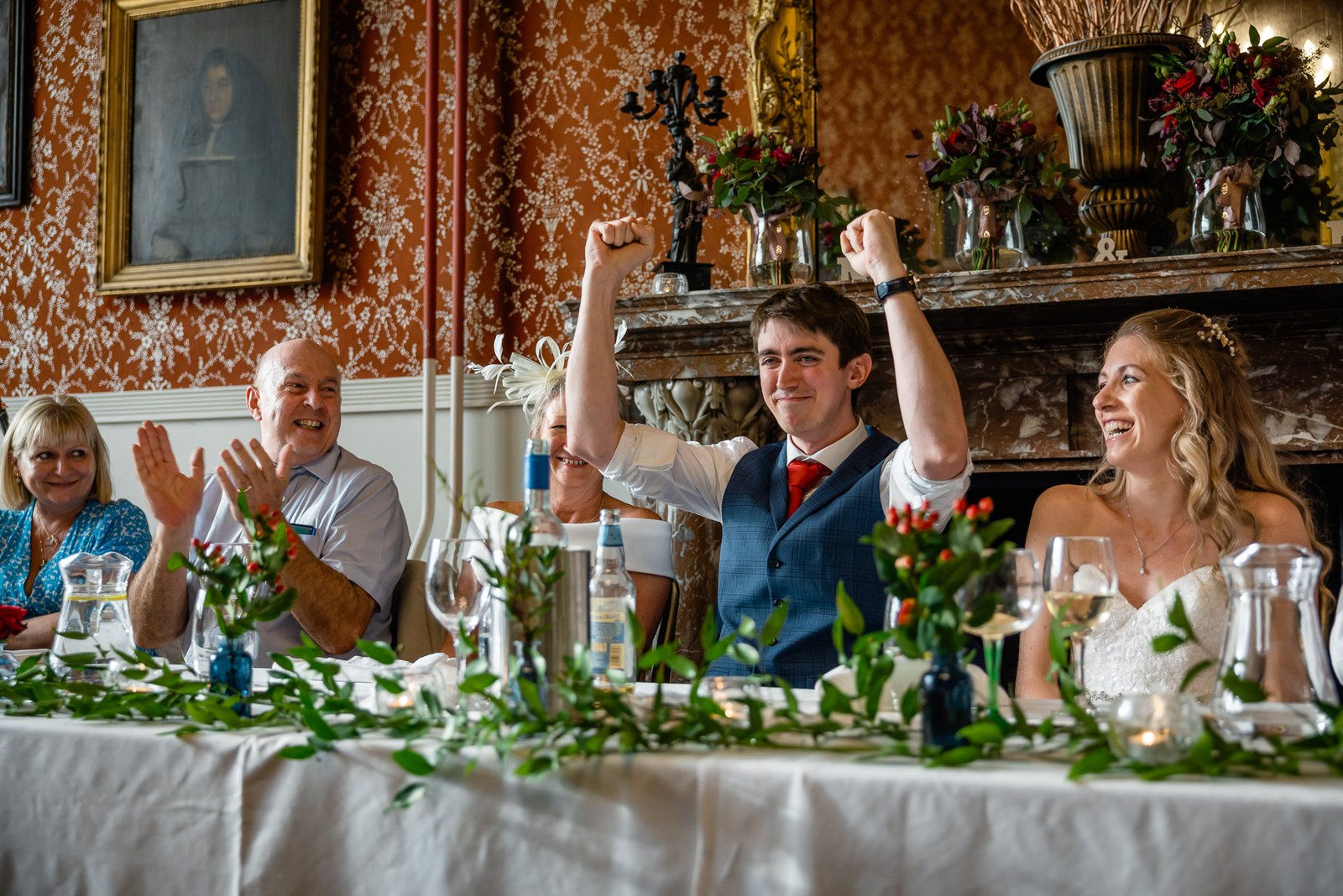 Groom cheering with raised arms at the top table as the bride laughs beside him