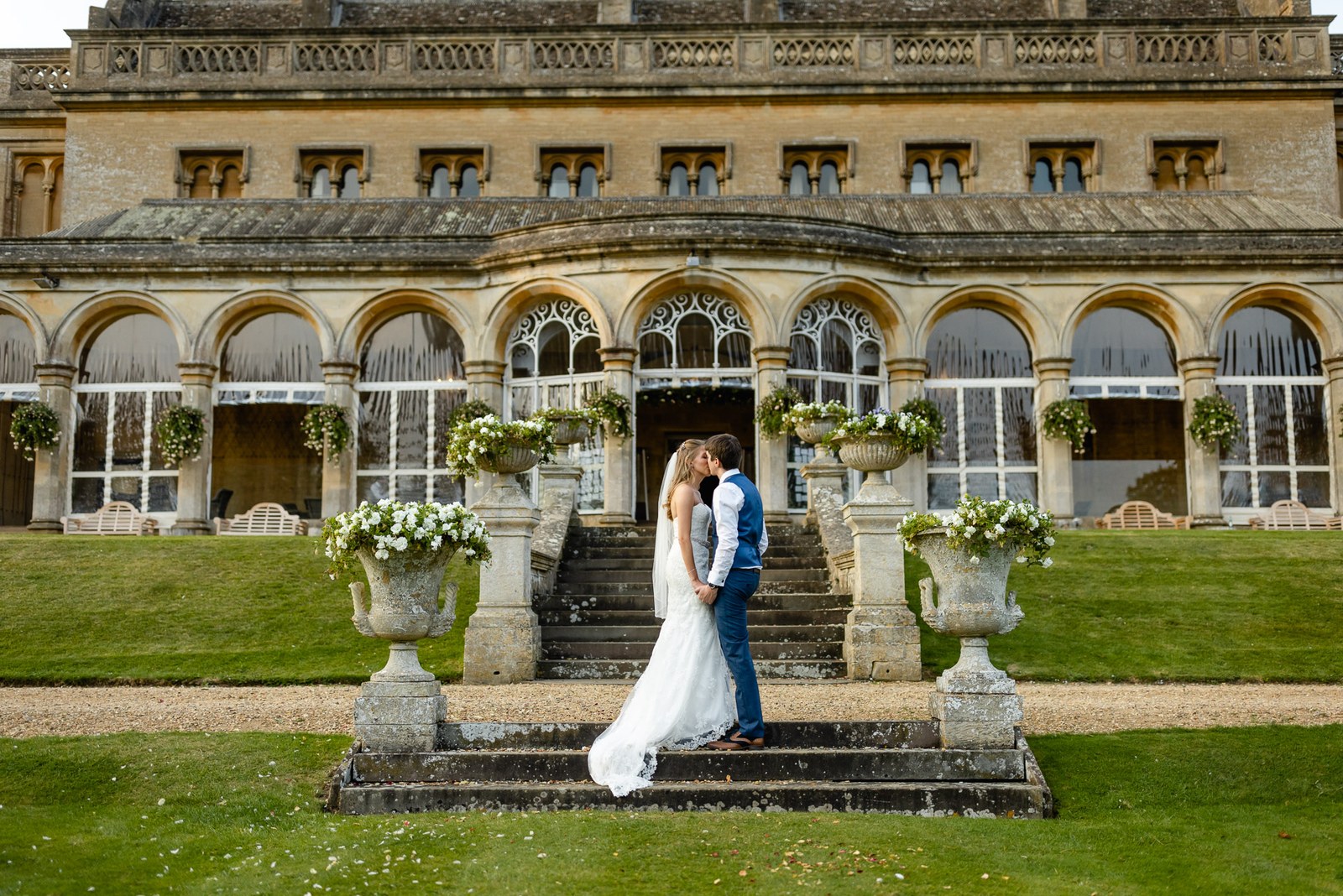 The couple between stone urns on the lawn before the honey-stone frontage of Grittleton House
