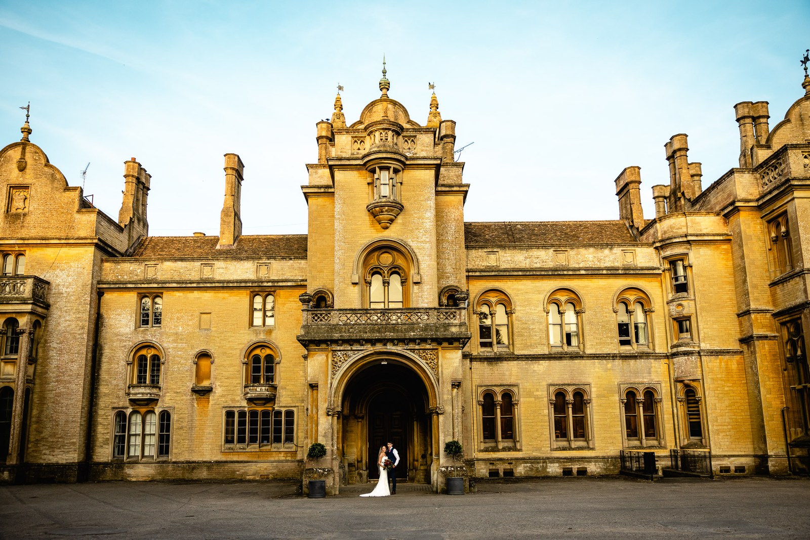 The golden Gothic frontage of Grittleton House glowing at sunset, the couple tiny before it