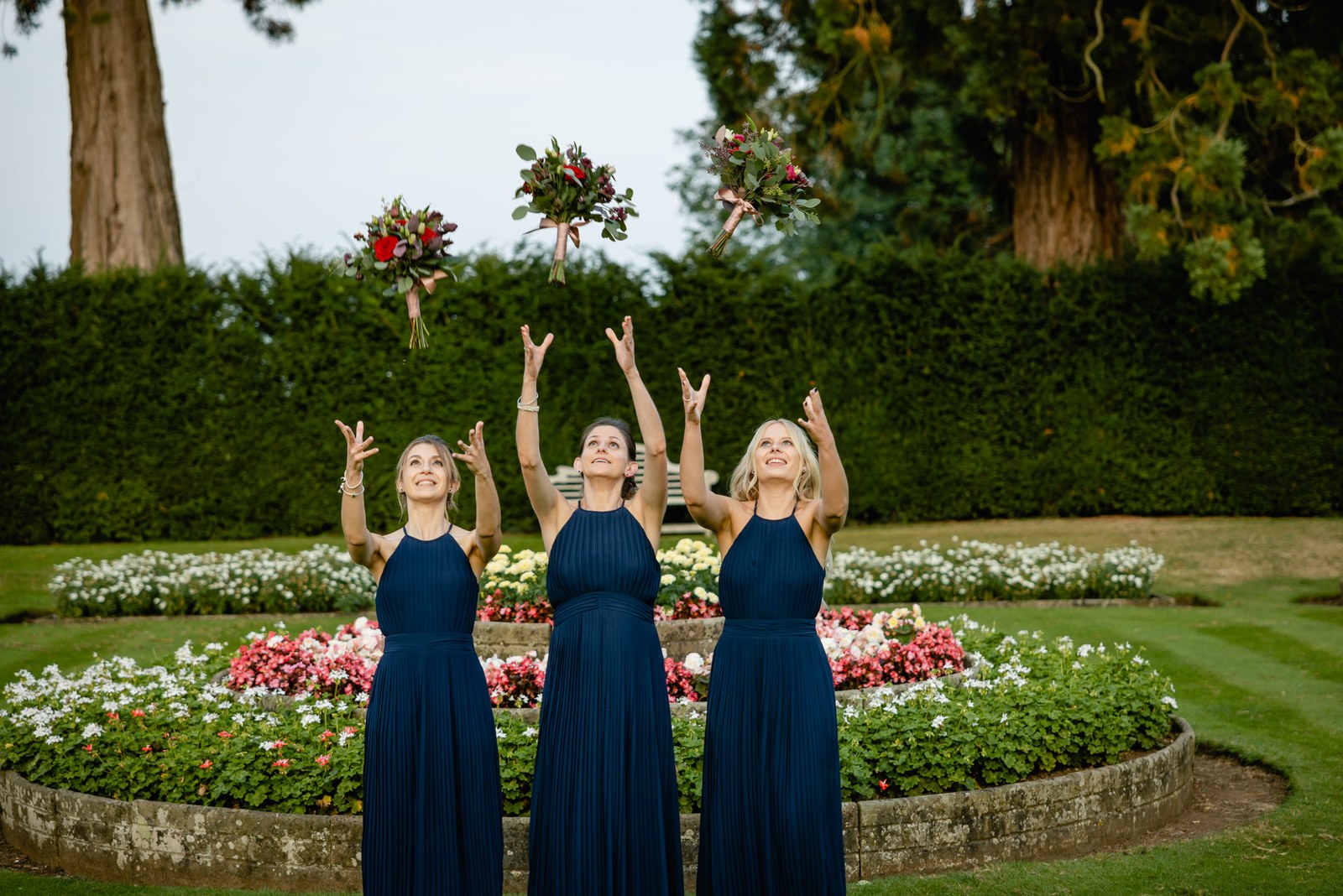 Bridesmaids laughing and tossing their bouquets skyward by the garden flower bed