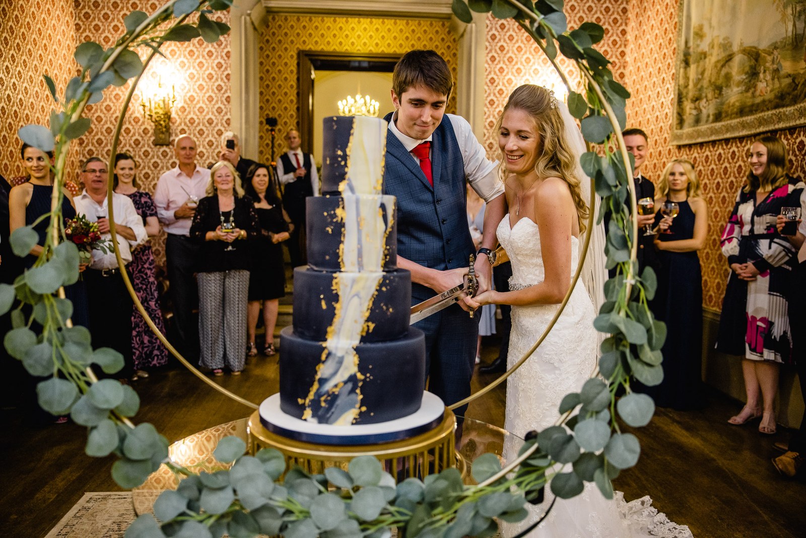 The couple cutting a navy-and-gold cake framed by a foliage hoop as guests watch
