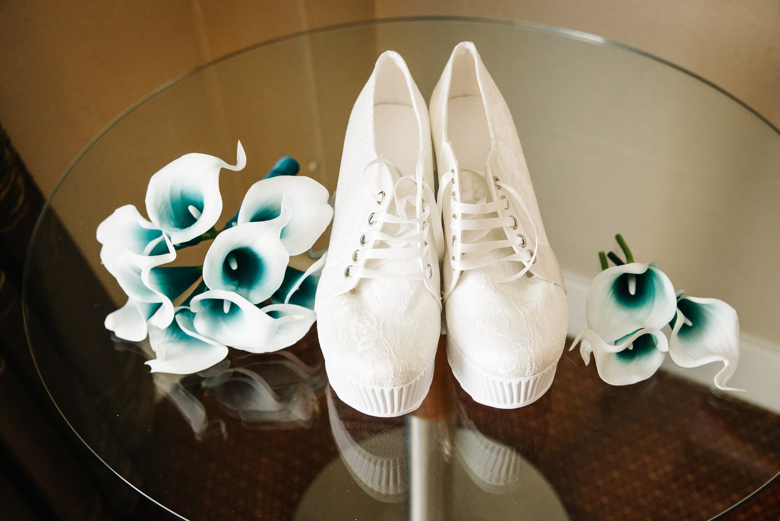 The bride's white shoes and teal calla lily bouquets arranged on a glass table