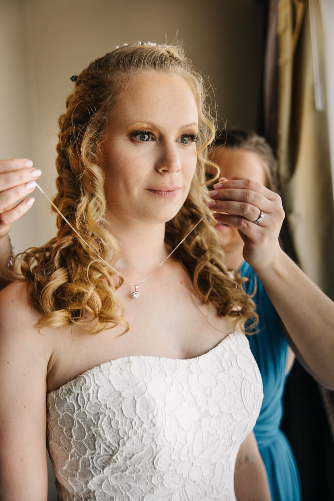The bride having her necklace fastened, a bridesmaid helping behind her