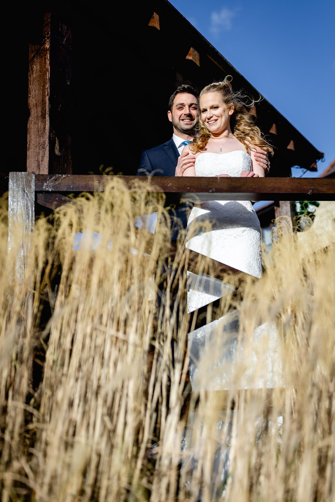 Low-angle shot of the newlyweds on a wooden balcony against a bright blue sky