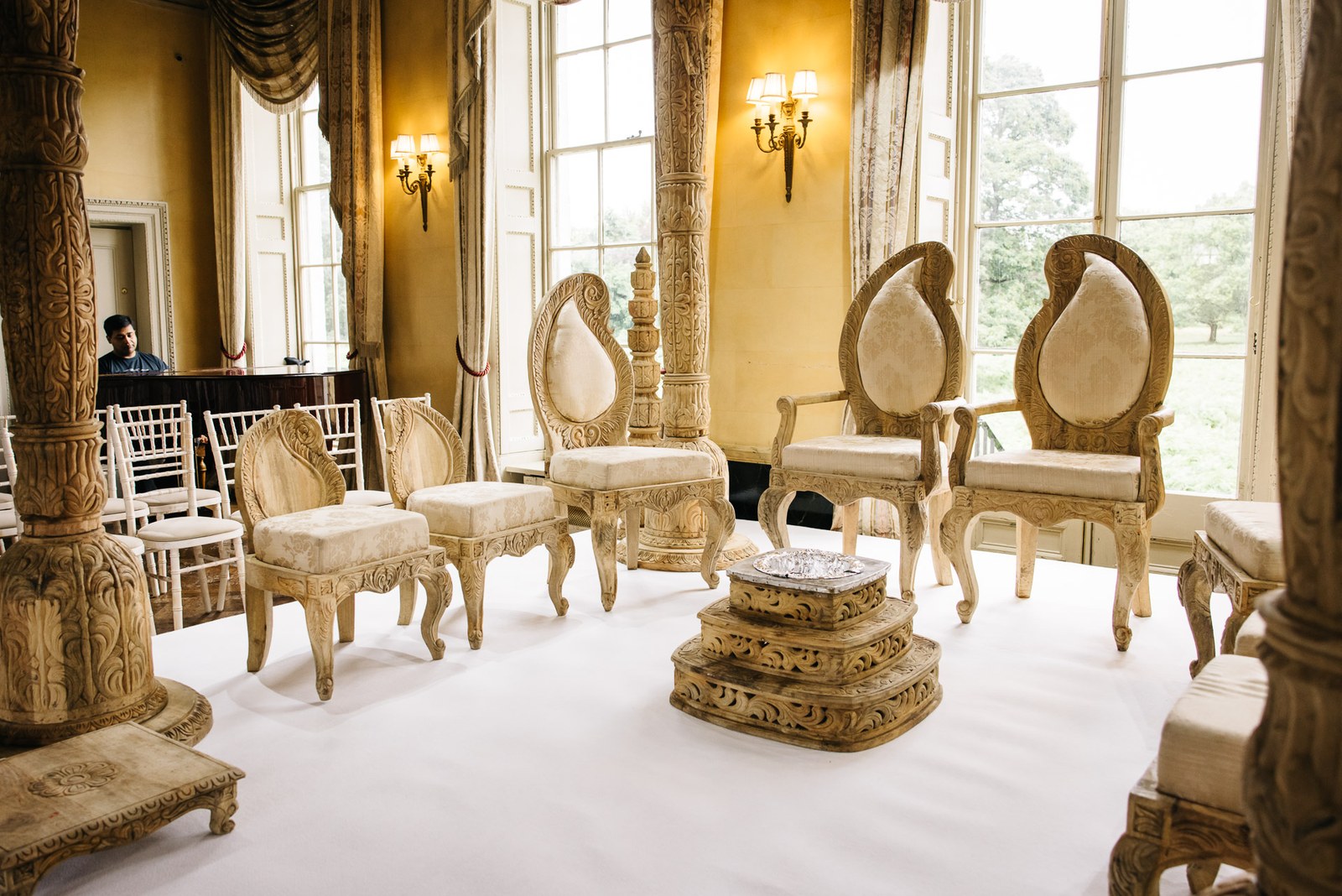 The ceremony room set with a carved mandap and thrones on a white-draped floor by tall windows