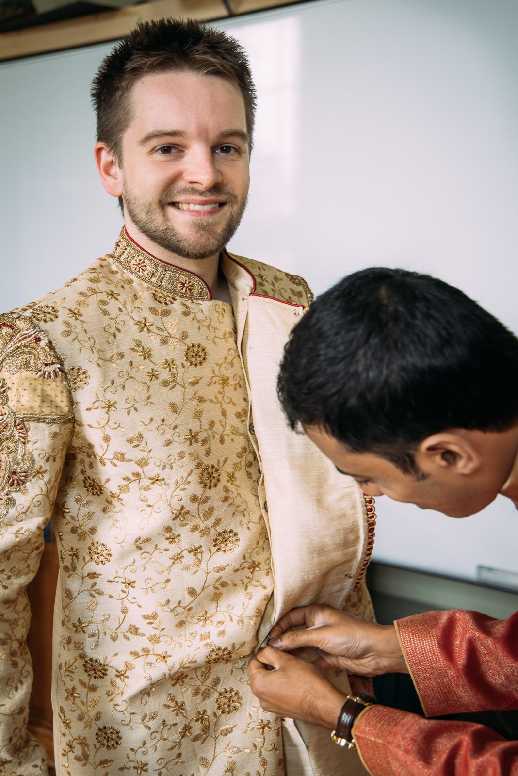 The groom in a cream-and-gold embroidered sherwani being helped to dress by an attendant