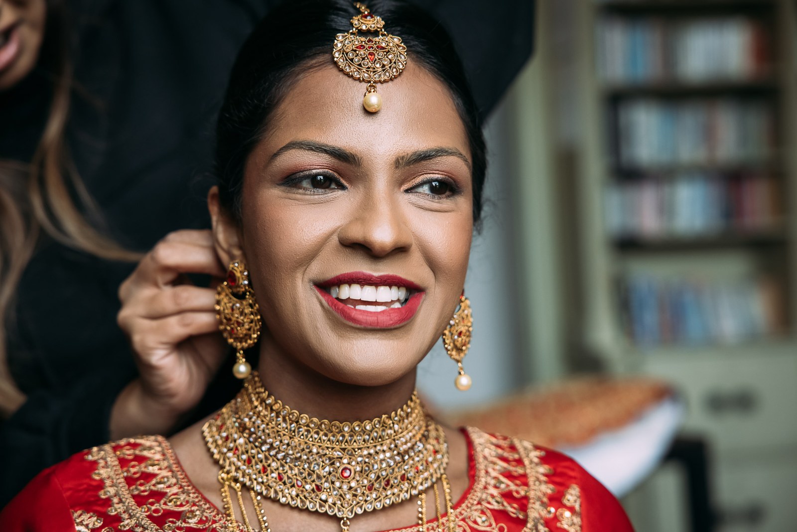 Bride smiling as an attendant fastens her earring, wearing gold jewellery and a maang tikka