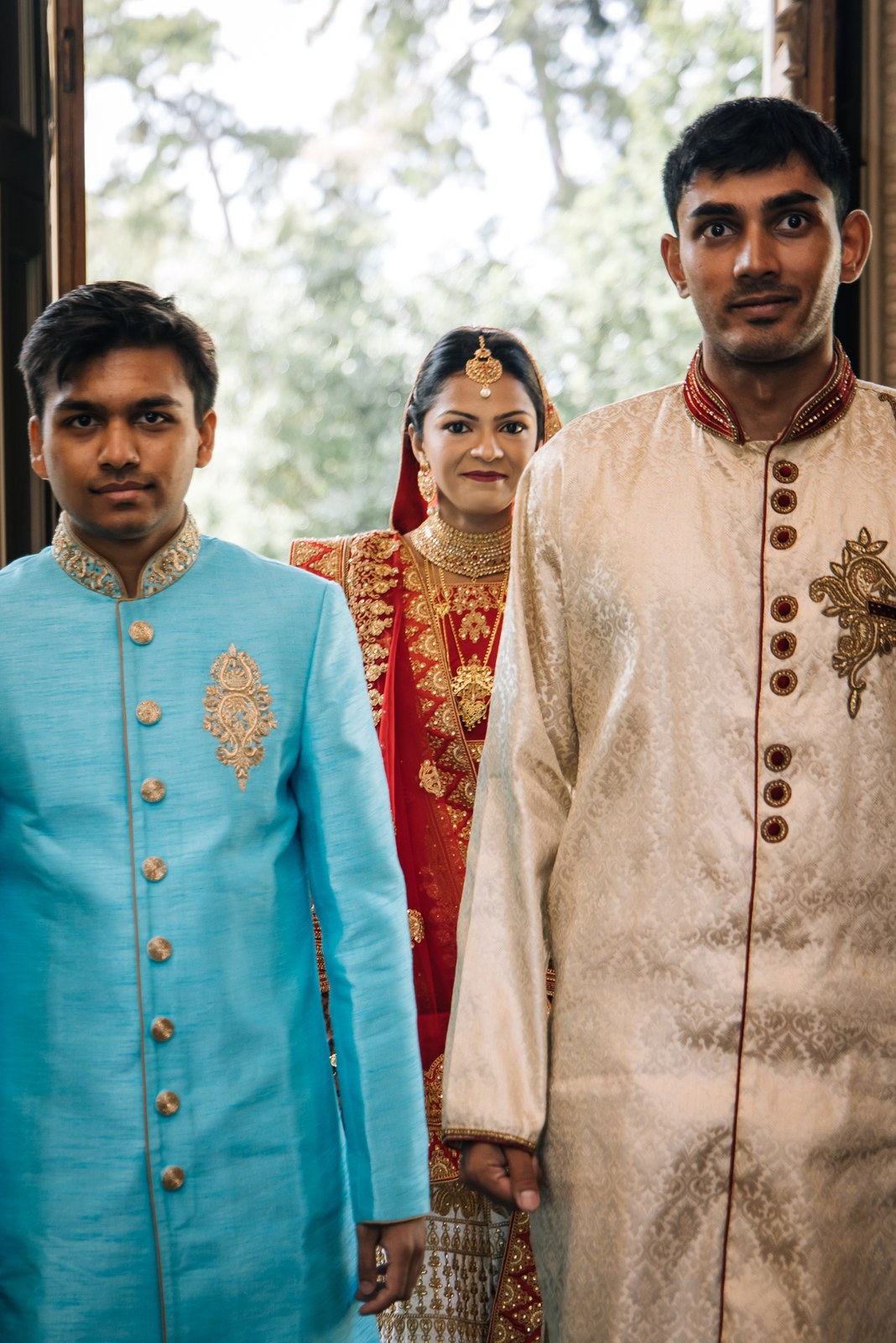 The bride flanked by two men in sherwanis in a sunlit doorway