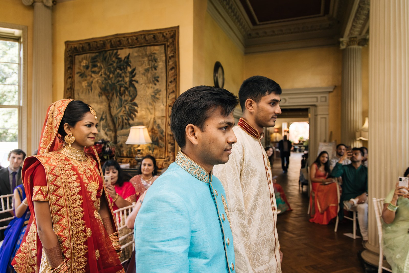 Bride escorted before seated guests beneath a tapestry in the ceremony room