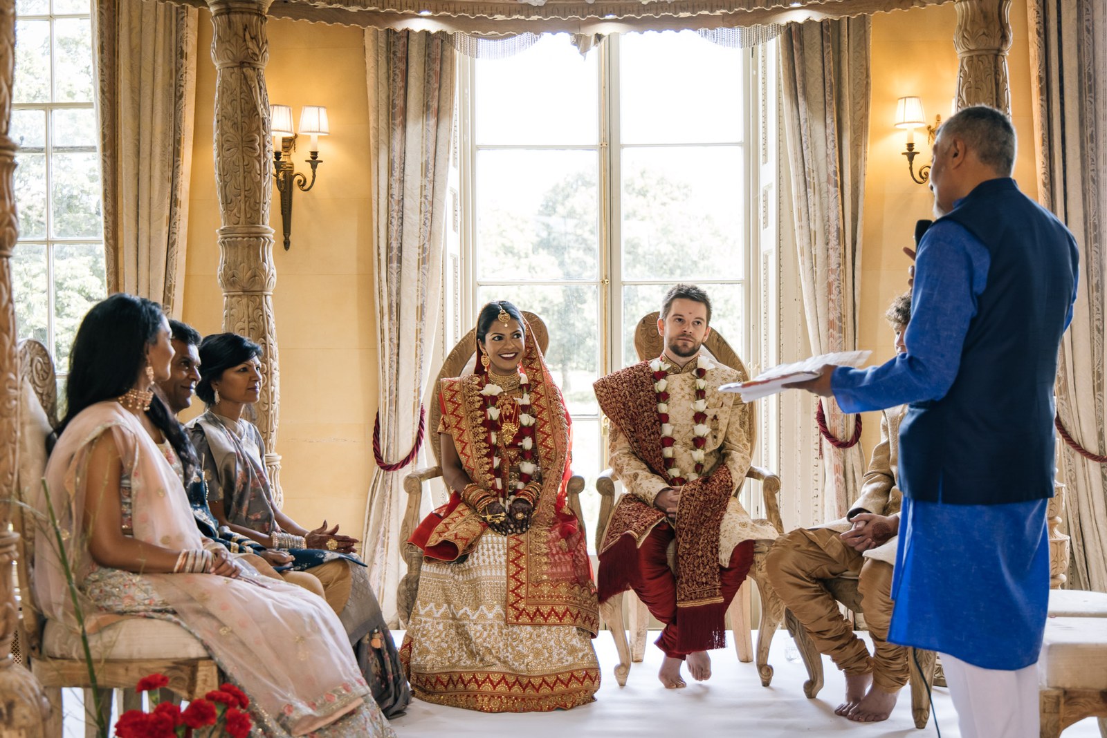Wide view of the seated couple as the priest leads the ceremony by the bay window