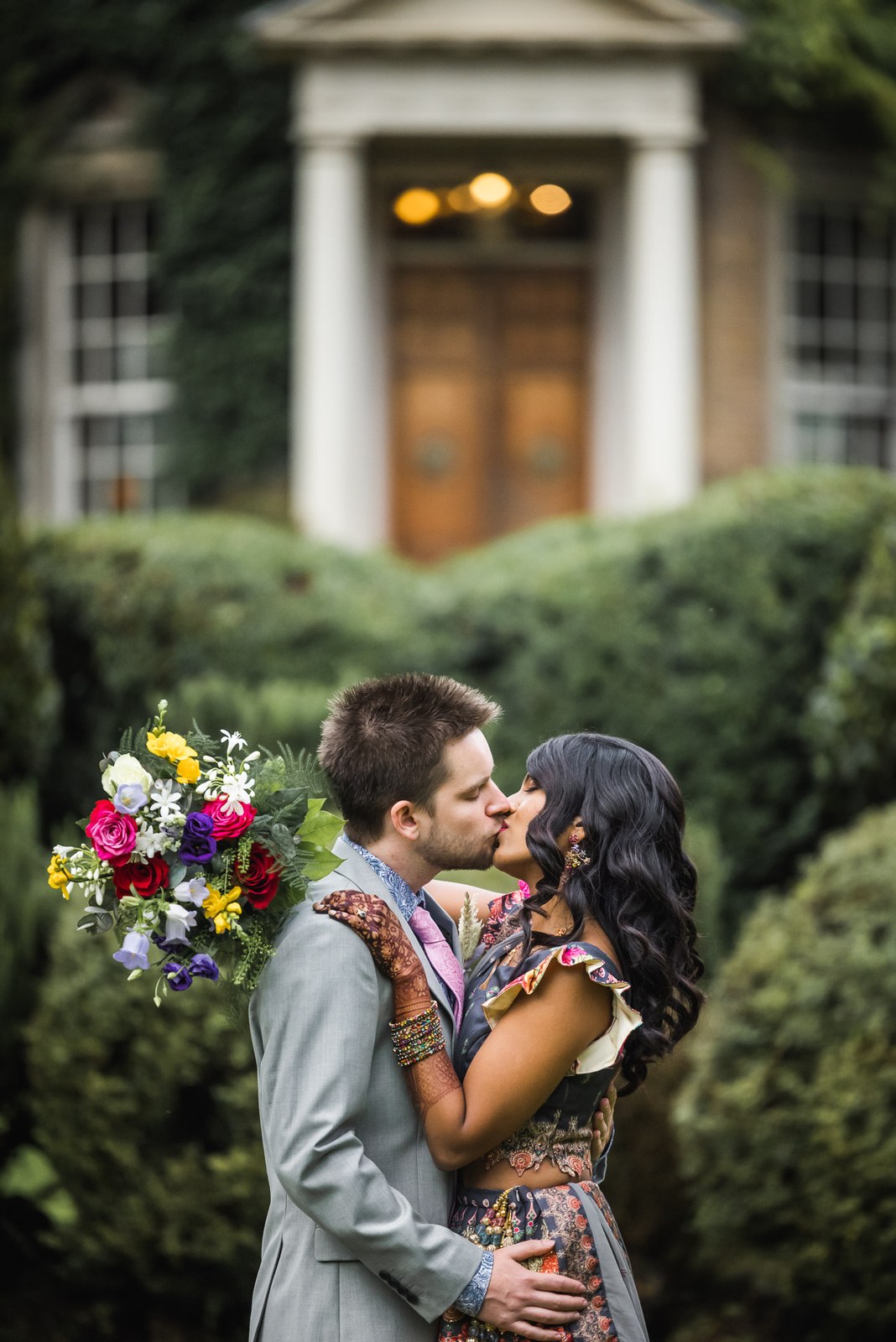 The couple kissing before the front door, framed by trimmed hedges