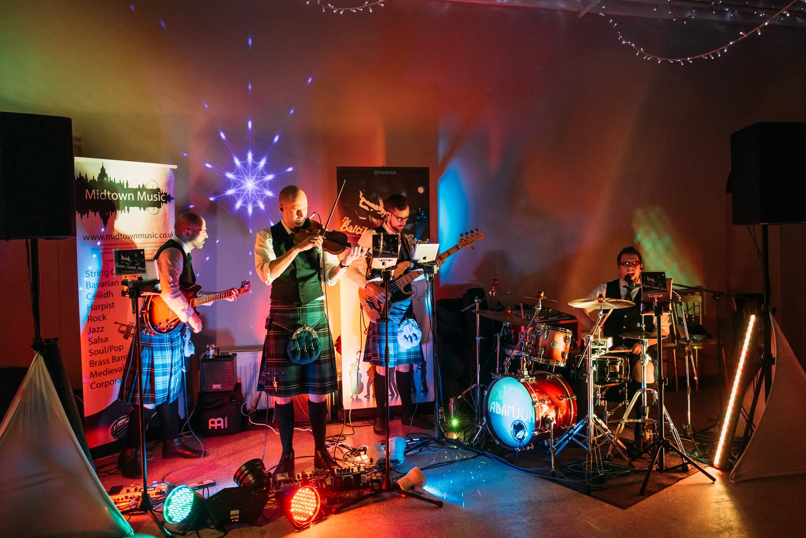 A kilted ceilidh band playing on stage under coloured lights