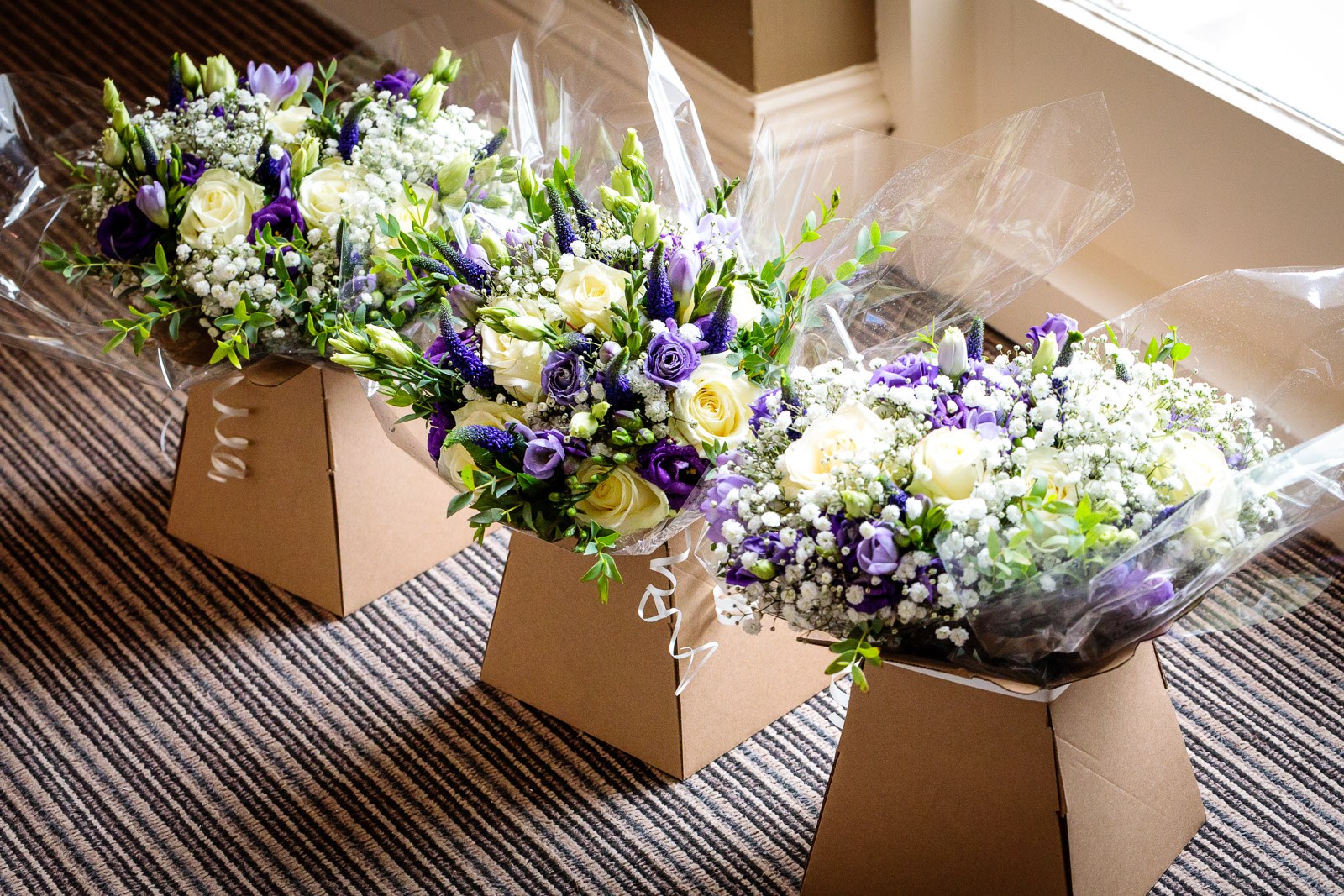Bridal bouquets of white roses and purple blooms resting in their delivery boxes on a striped carpet