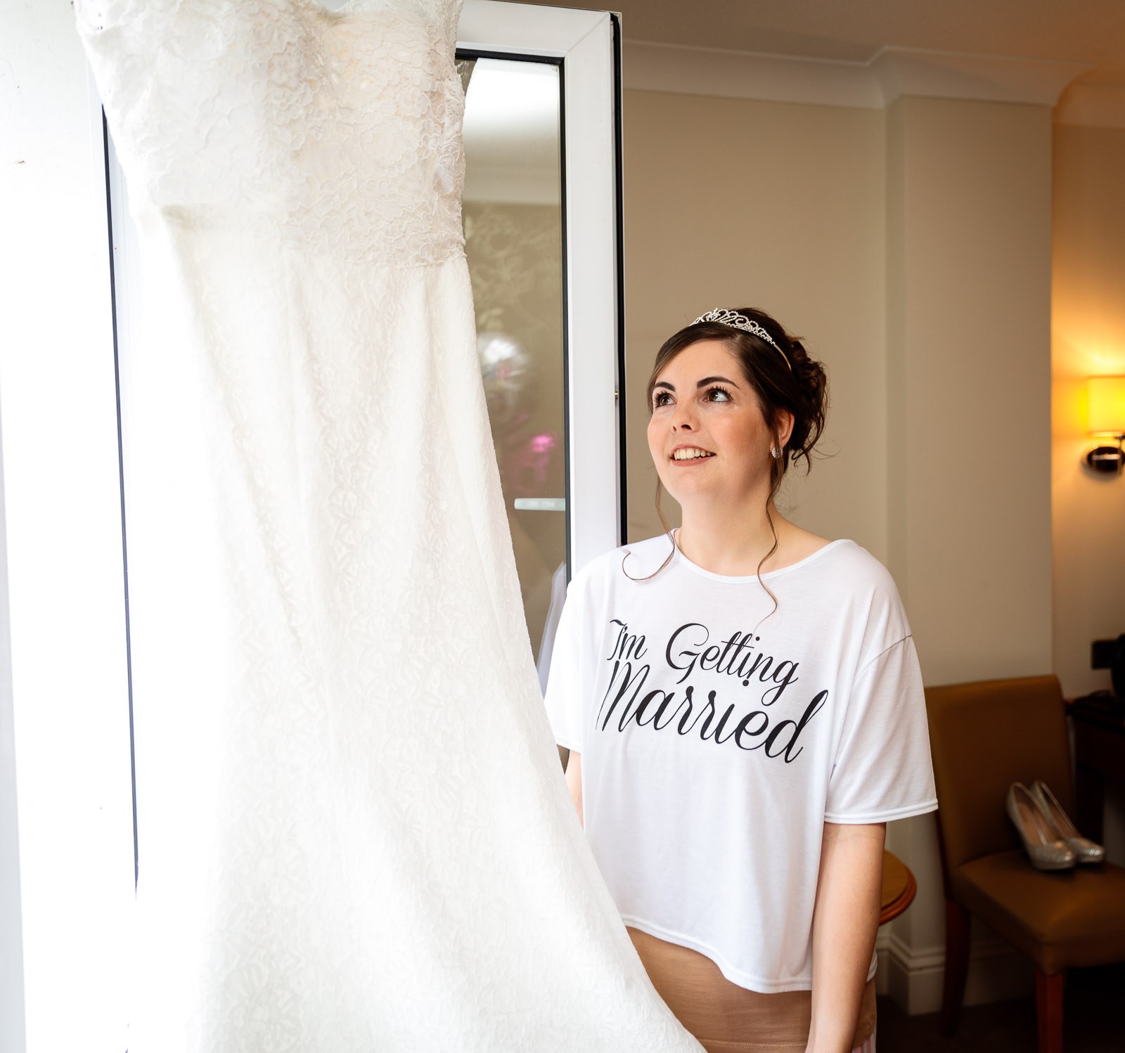 Bride in a slogan tee gazing up at her gown hanging in the window light