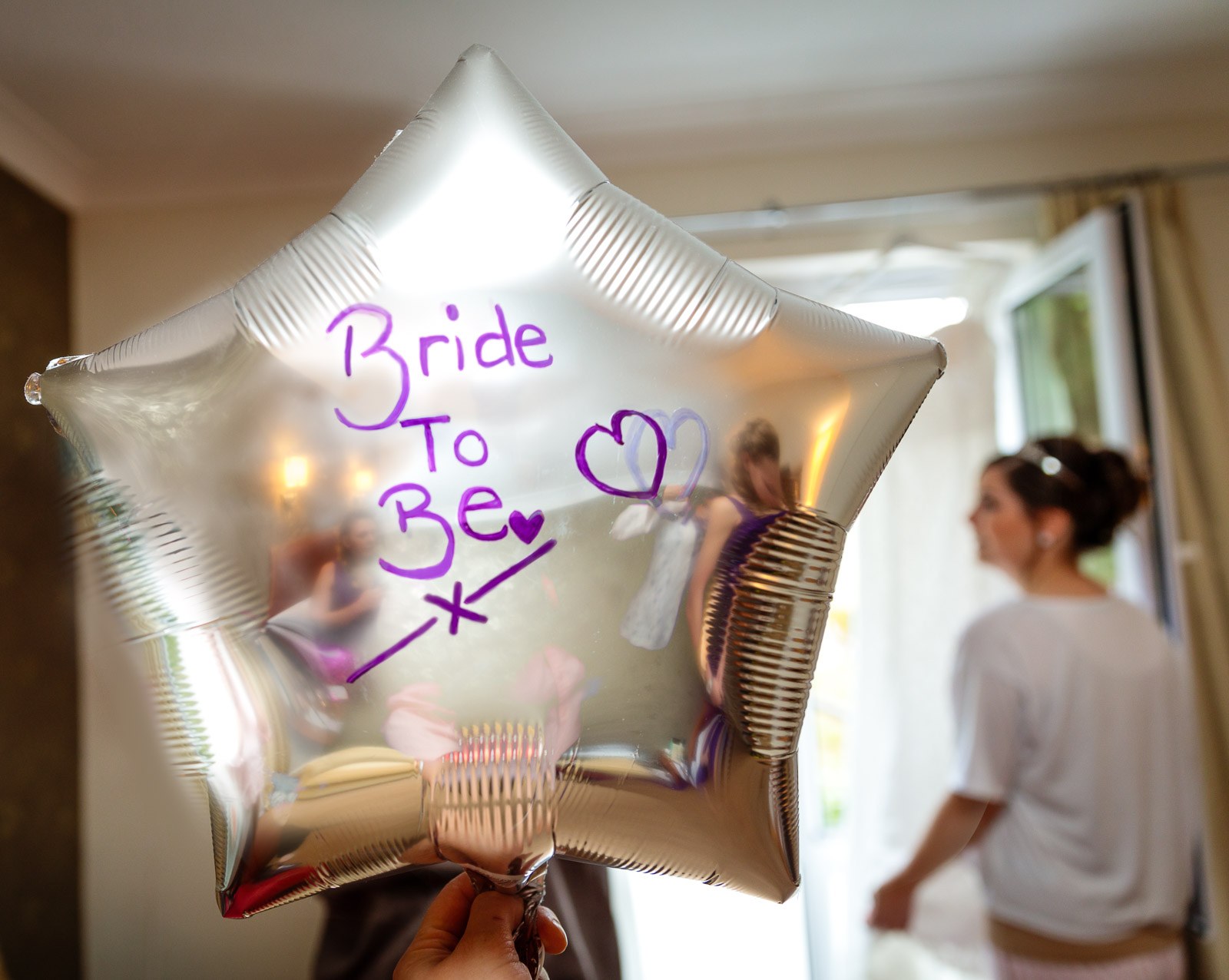 A hand holding a silver 'Bride To Be' star balloon as the bride prepares behind