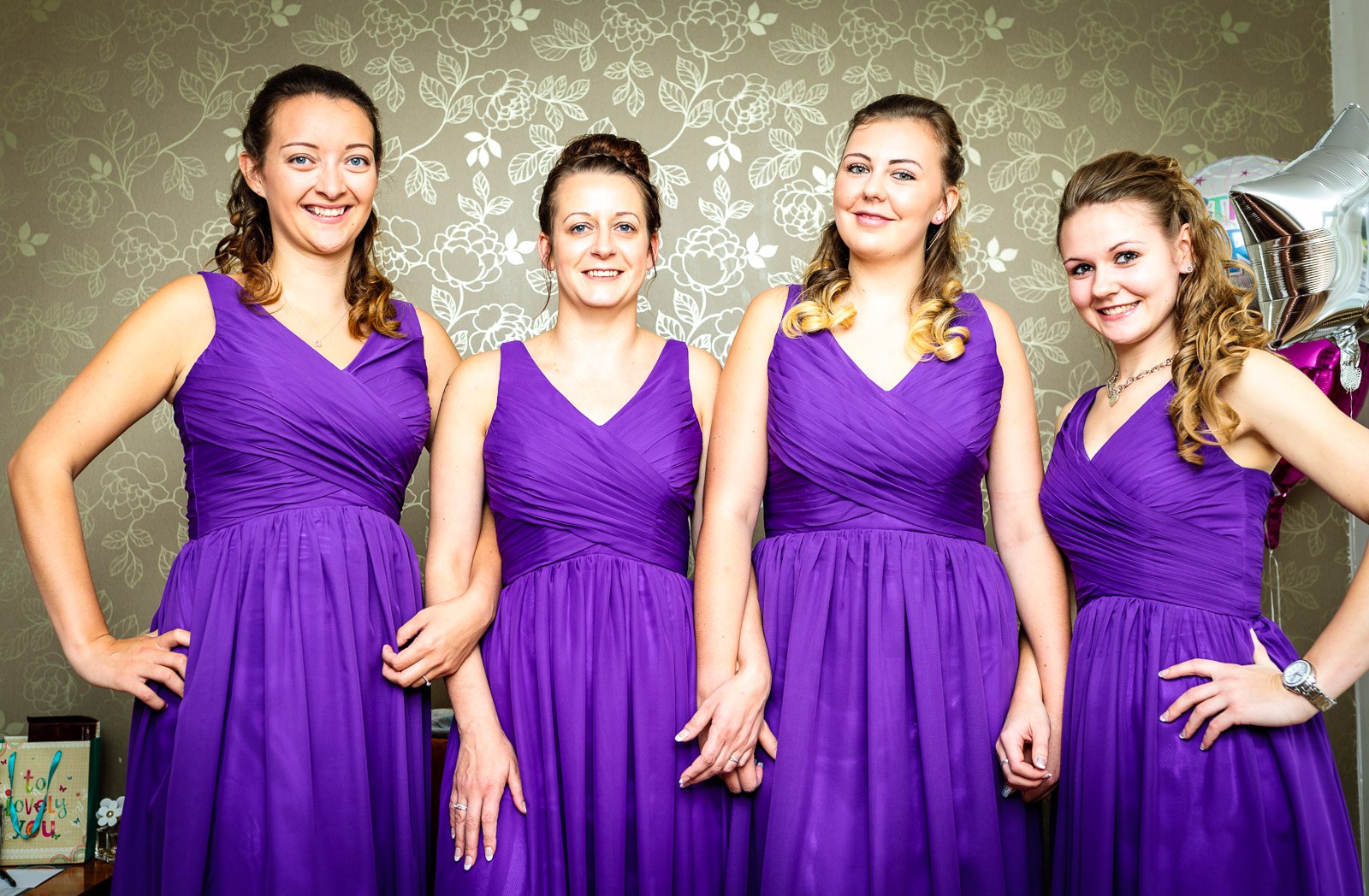 Four bridesmaids in flowing purple gowns posed against floral wallpaper
