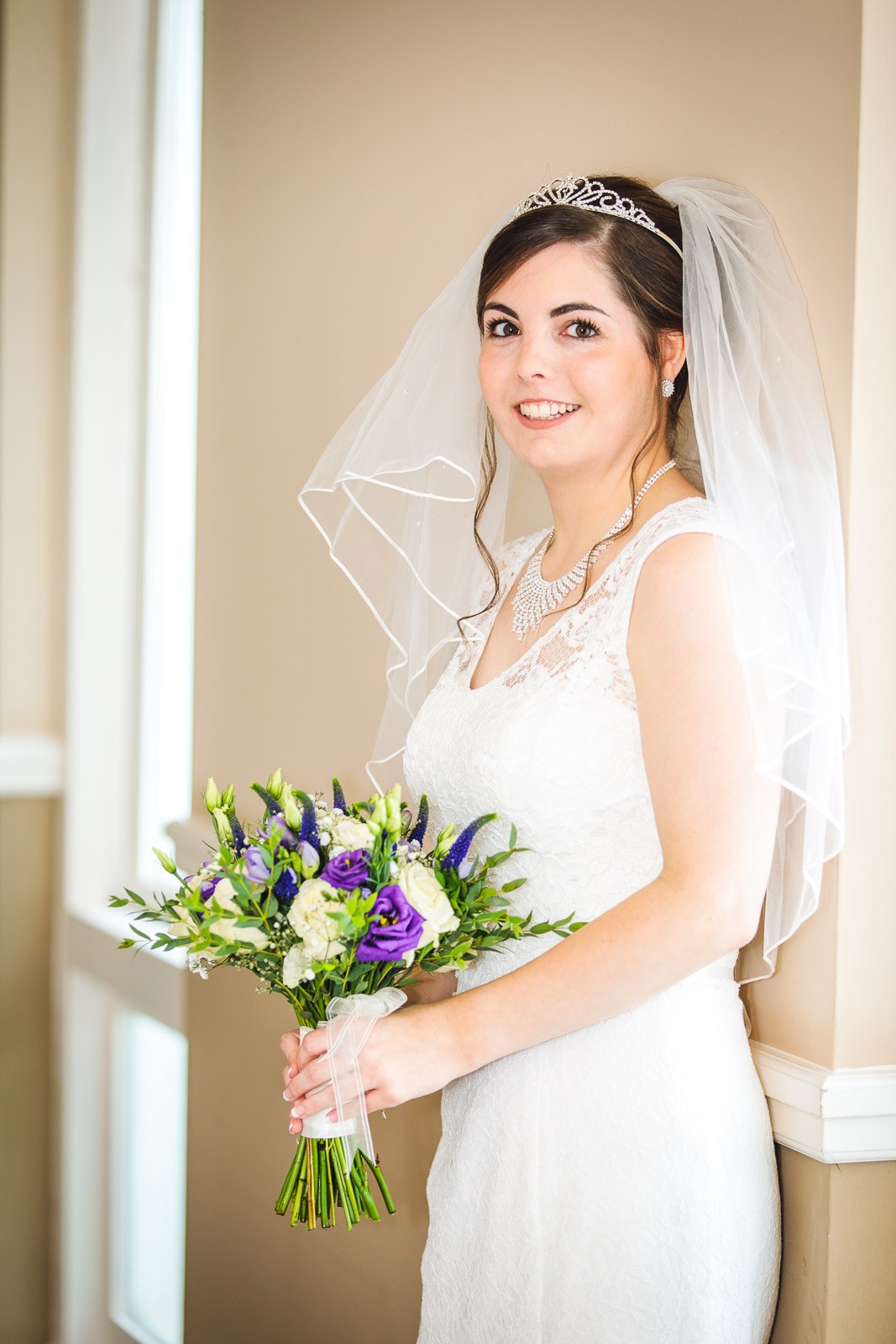 Bride holding her purple-and-white bouquet in soft window light
