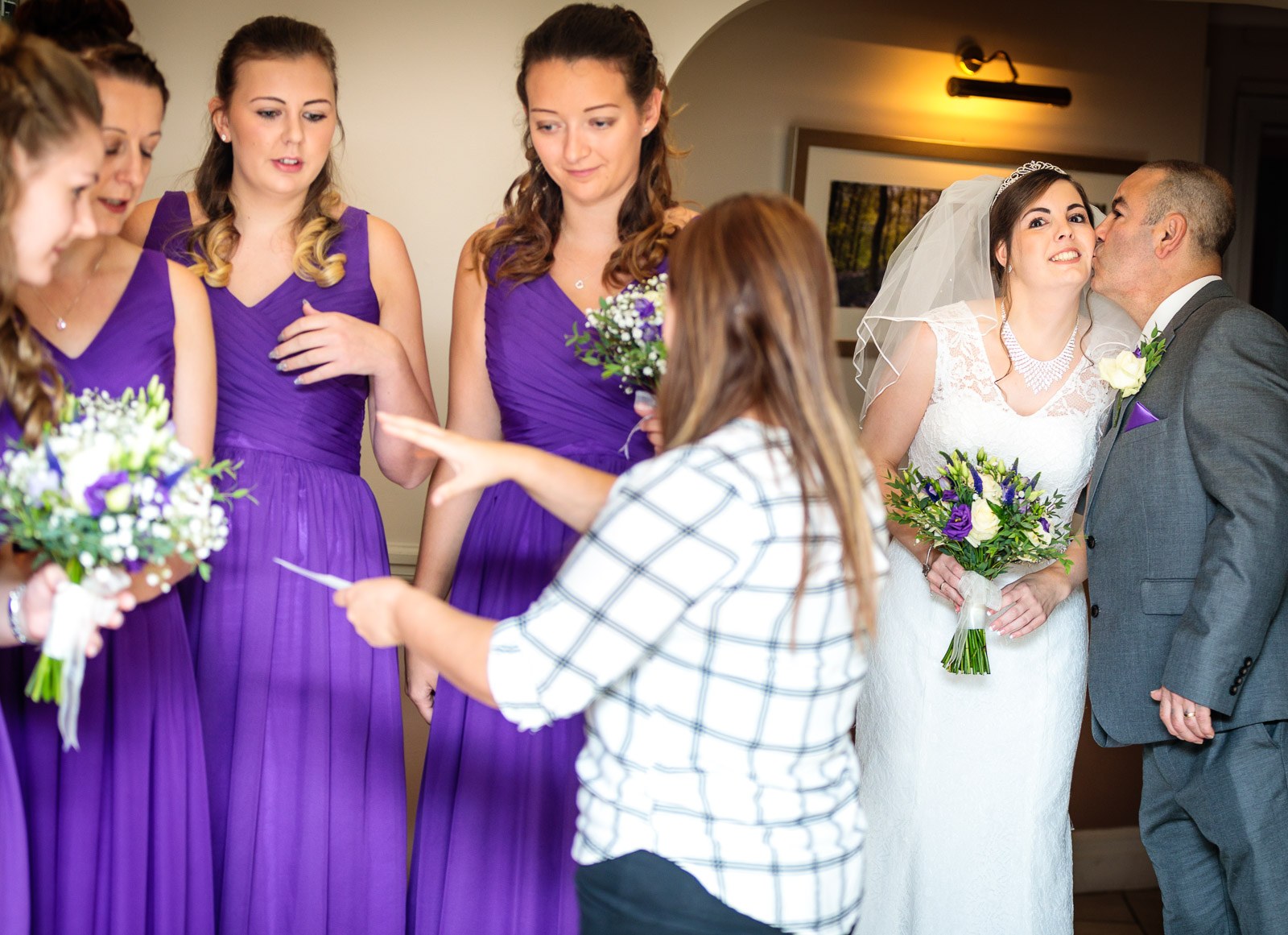 The father of the bride kissing her cheek as the bridesmaids wait with their bouquets