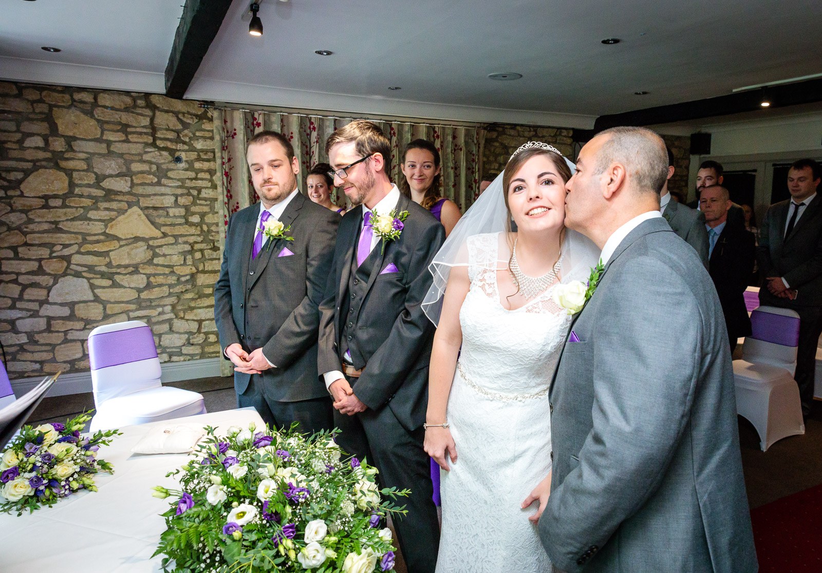 The father of the bride kissing her at the ceremony against an exposed stone wall