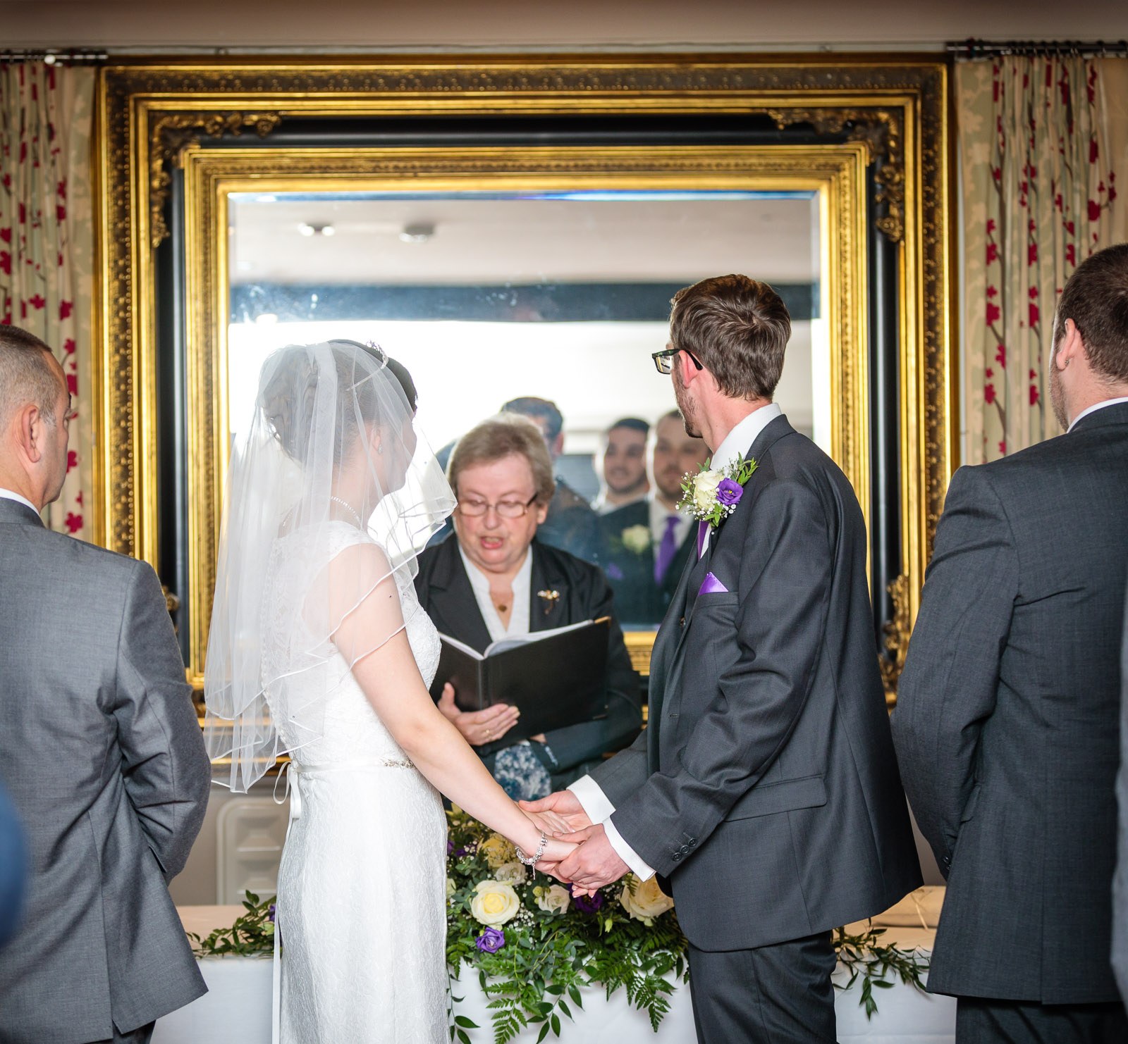 The couple holding hands before the registrar, reflected in a gilt mirror