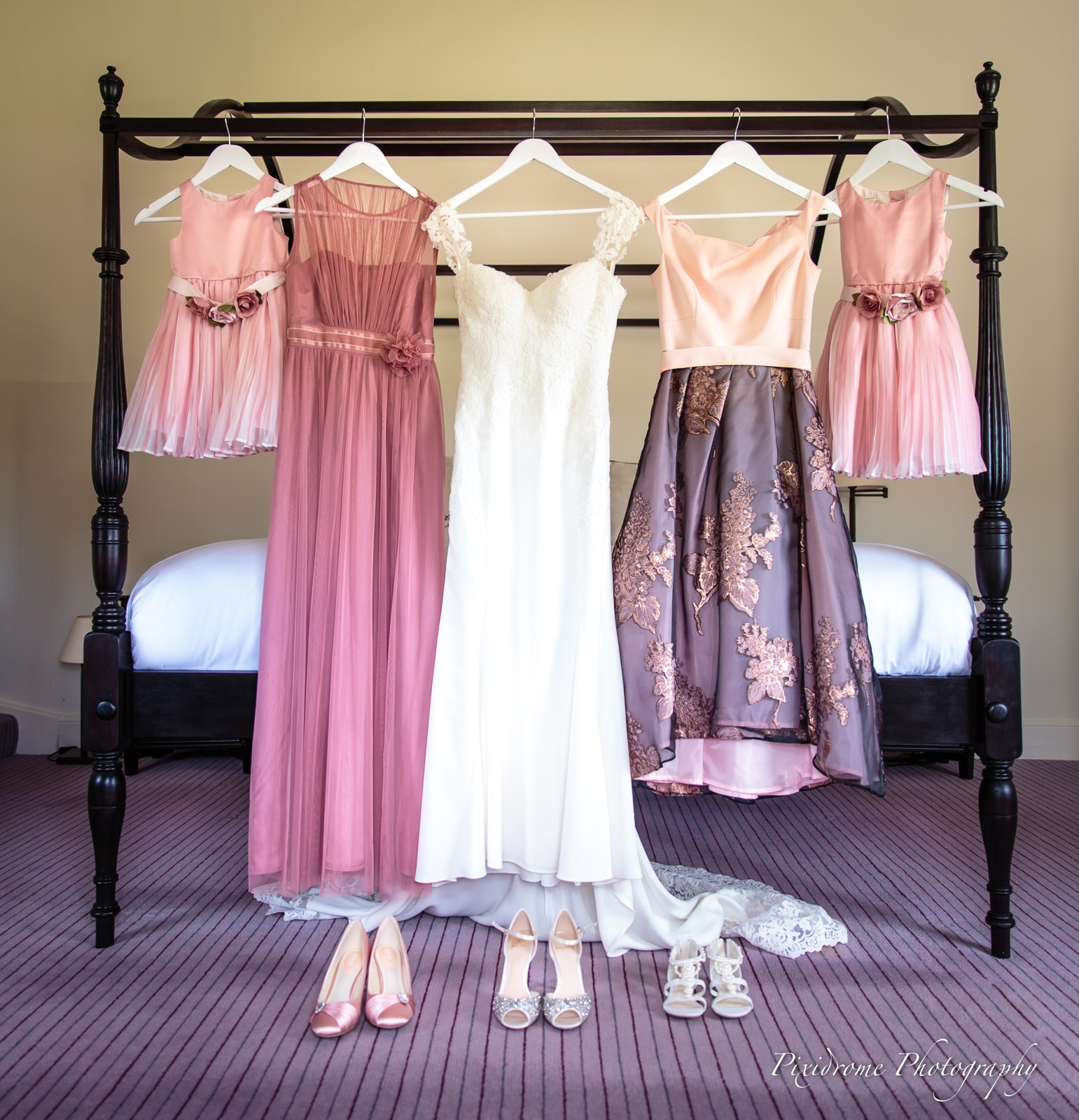 The wedding gown and pink bridesmaid dresses hanging on a four-poster bed, shoes lined up below