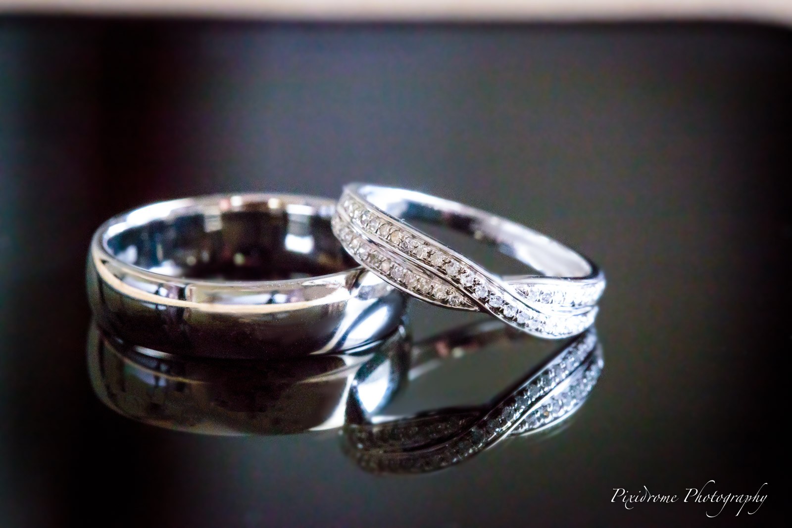Close-up of the two wedding rings, one diamond-set, reflected on a dark surface