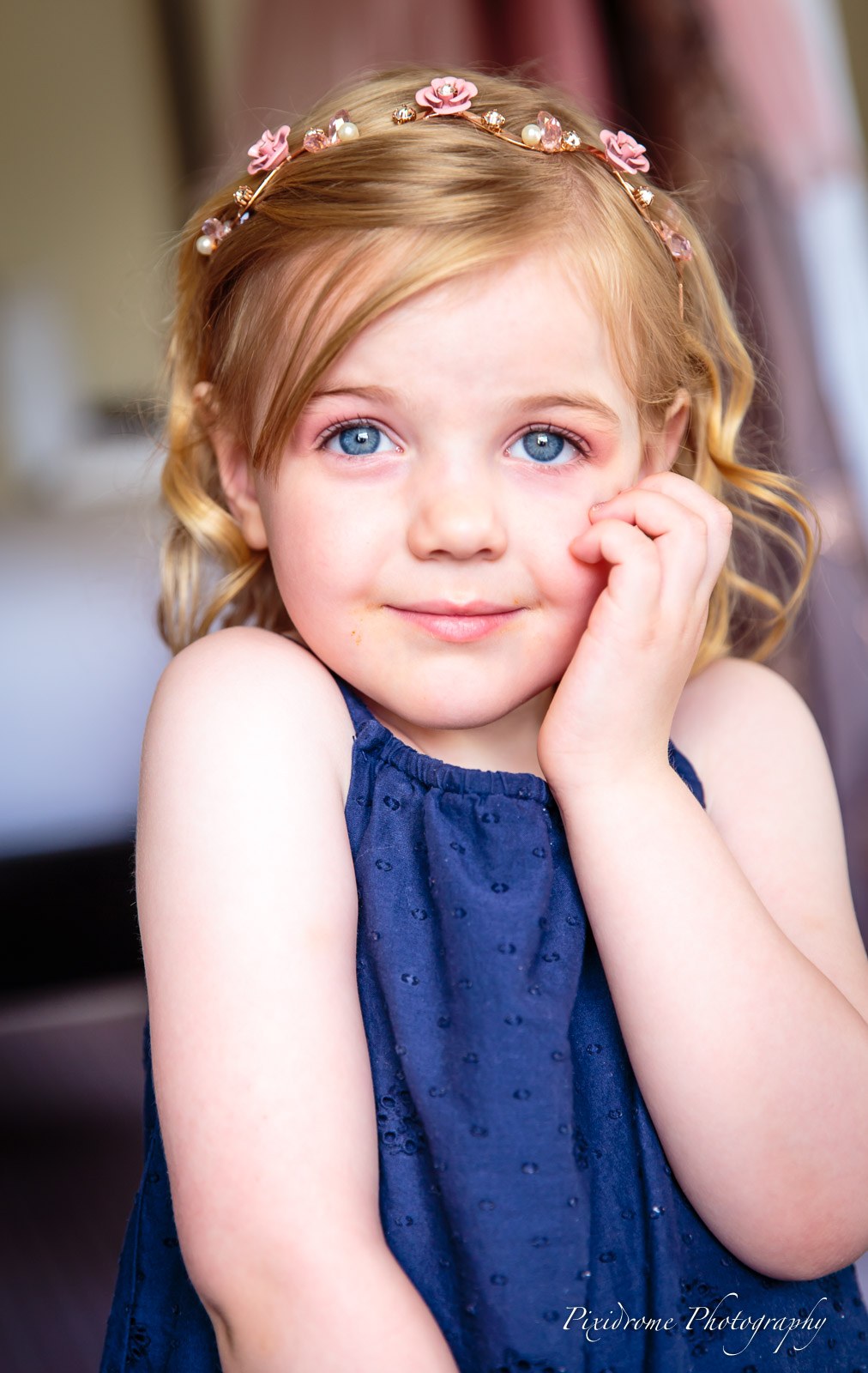 A young flower girl in a navy dress and pink floral headband resting her chin on her hand