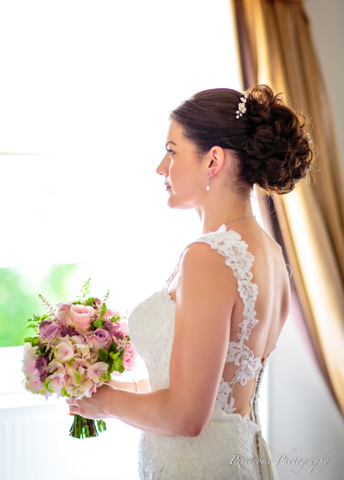 Bride at the window with her bouquet, the open lace back of her gown catching the light