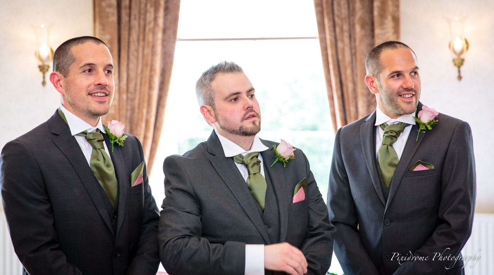 The groom and two groomsmen in grey suits and sage ties waiting by the window