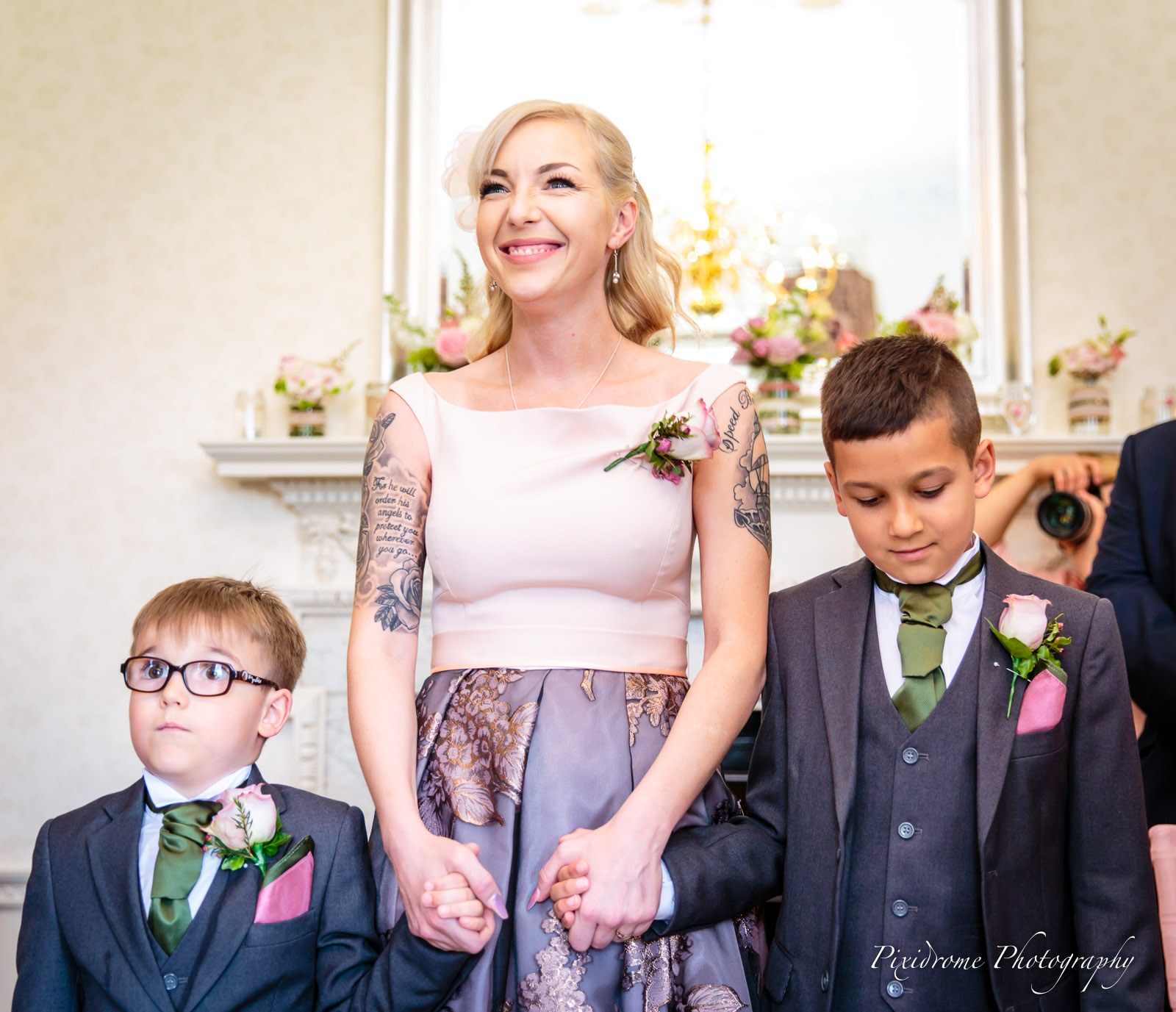 A woman in blush pink holding hands with two page boys at the ceremony