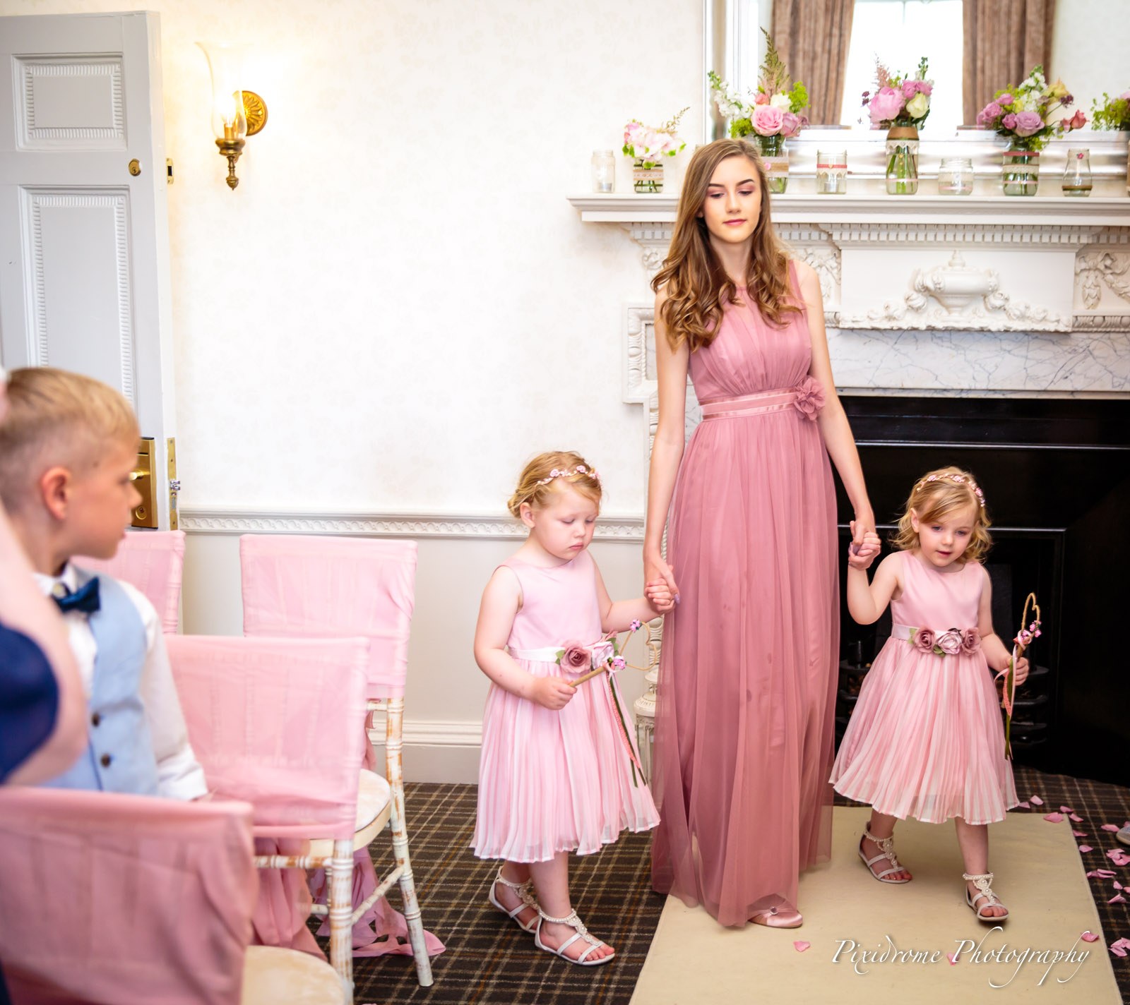 A bridesmaid leading the flower girls up the aisle past a flower-topped fireplace