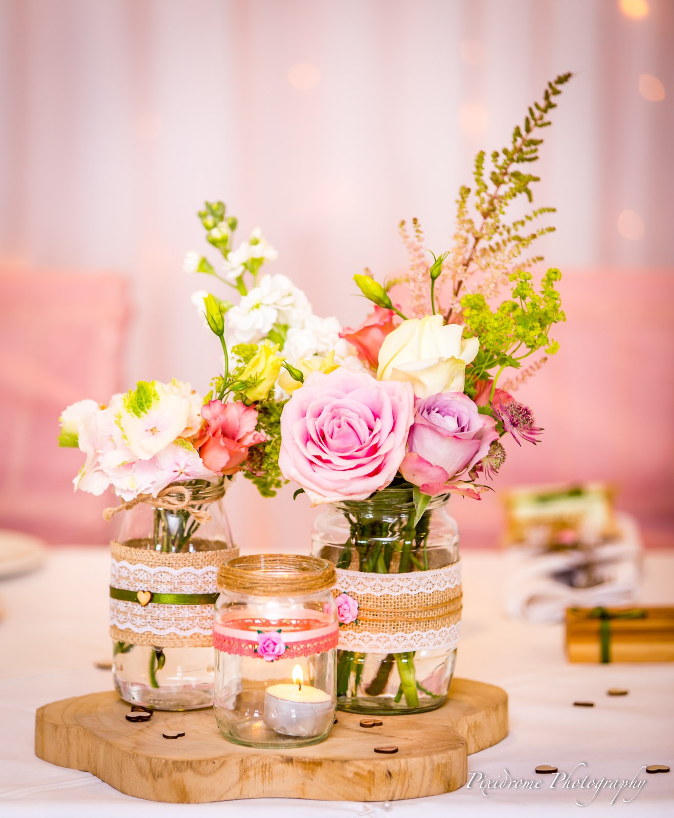 A table centrepiece of jam jars filled with pink roses beside a lit candle