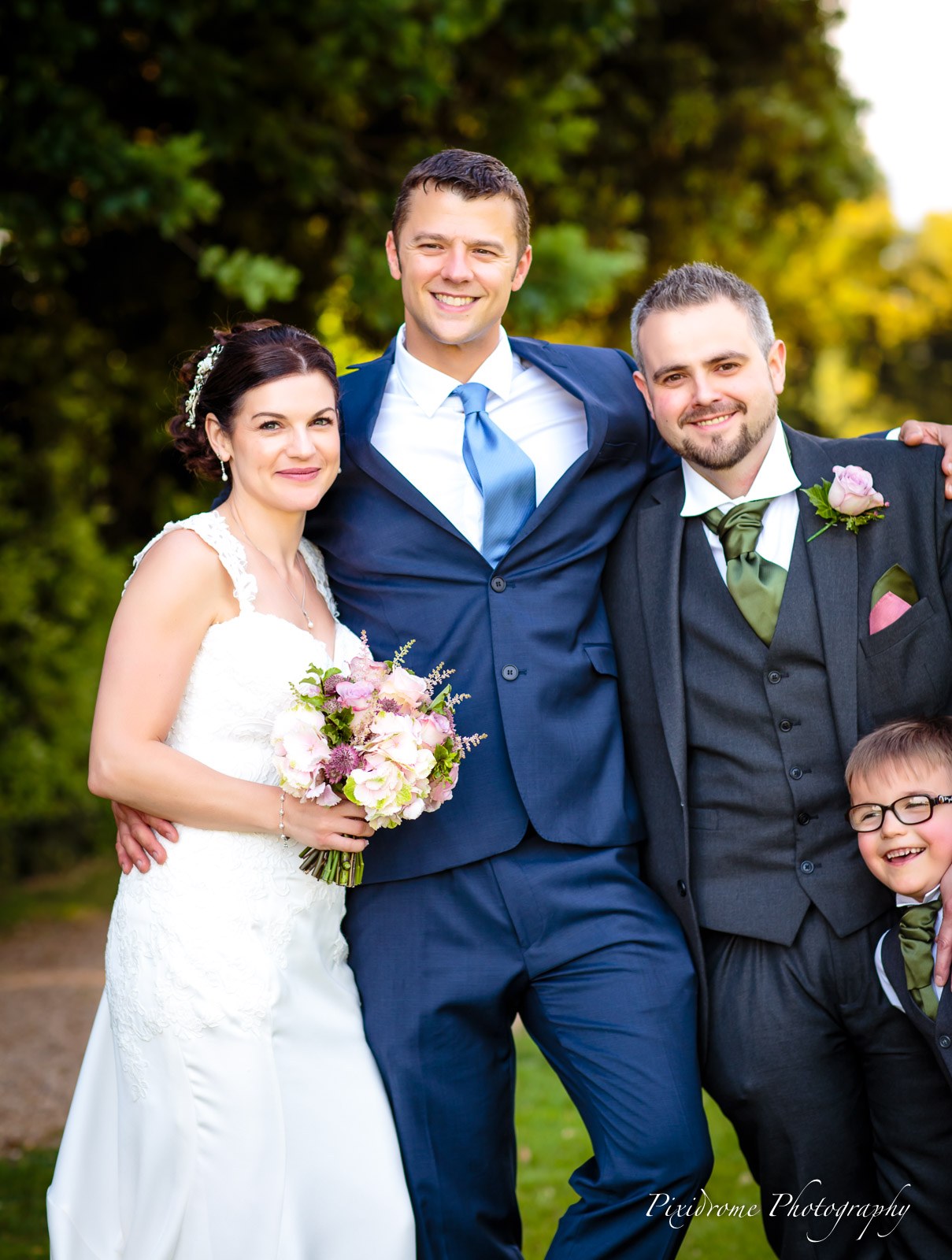 The couple and a guest in a blue suit by the hedge, a page boy peeking from behind