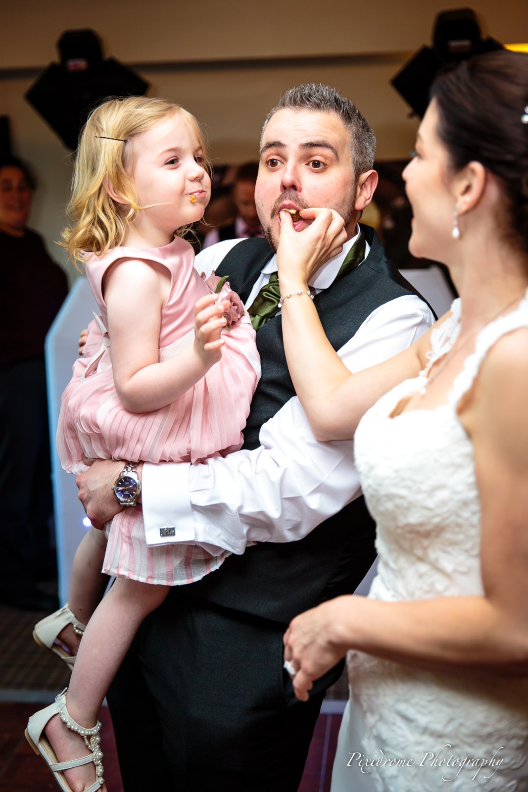 The groom holding a flower girl as the bride looks on fondly