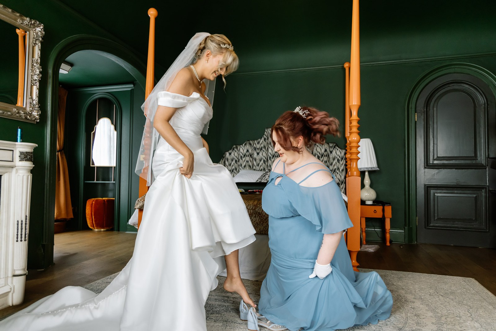A bridesmaid kneeling to adjust the bride's dress in the green four-poster room