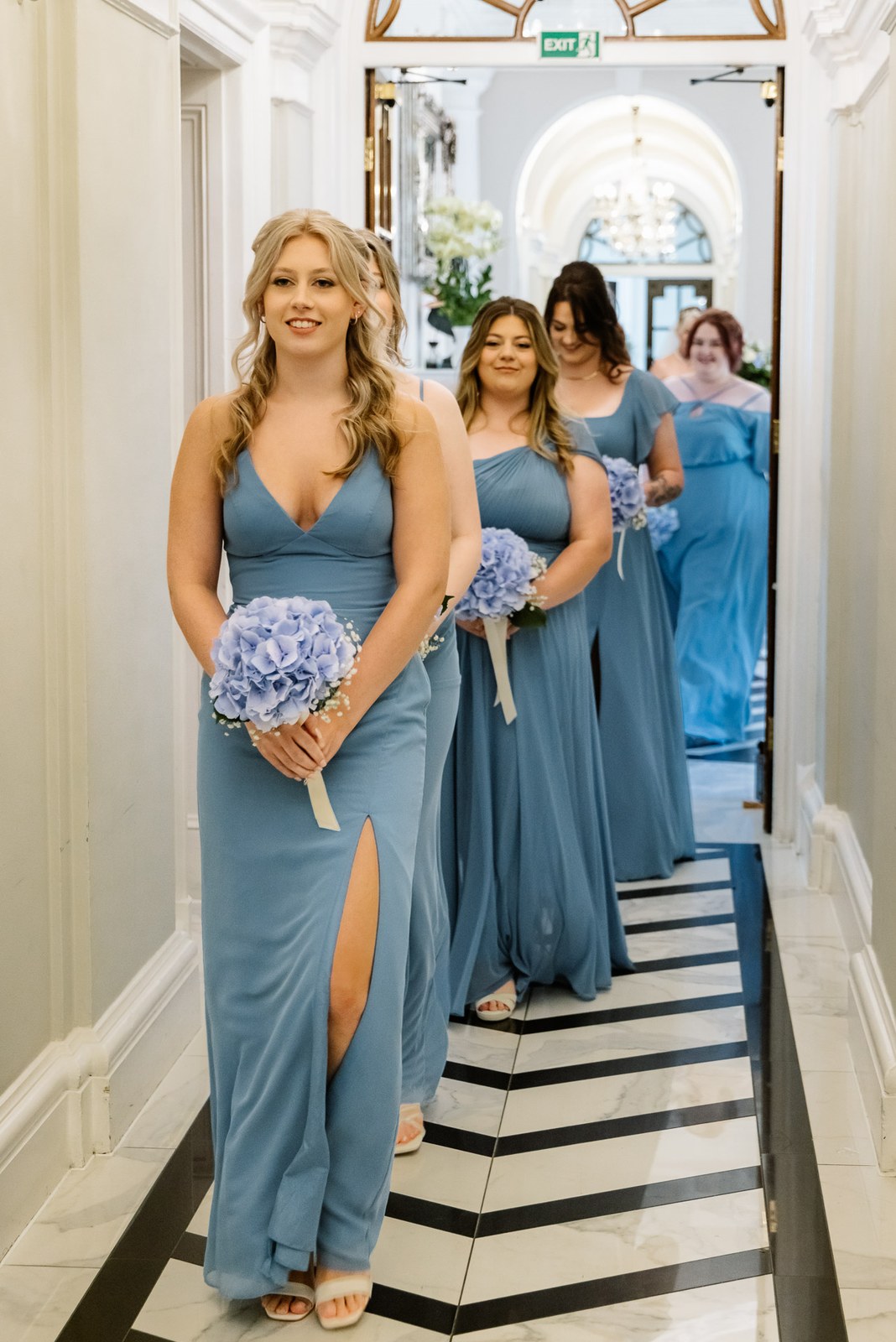 Bridesmaids carrying hydrangea bouquets down the striped-floor corridor
